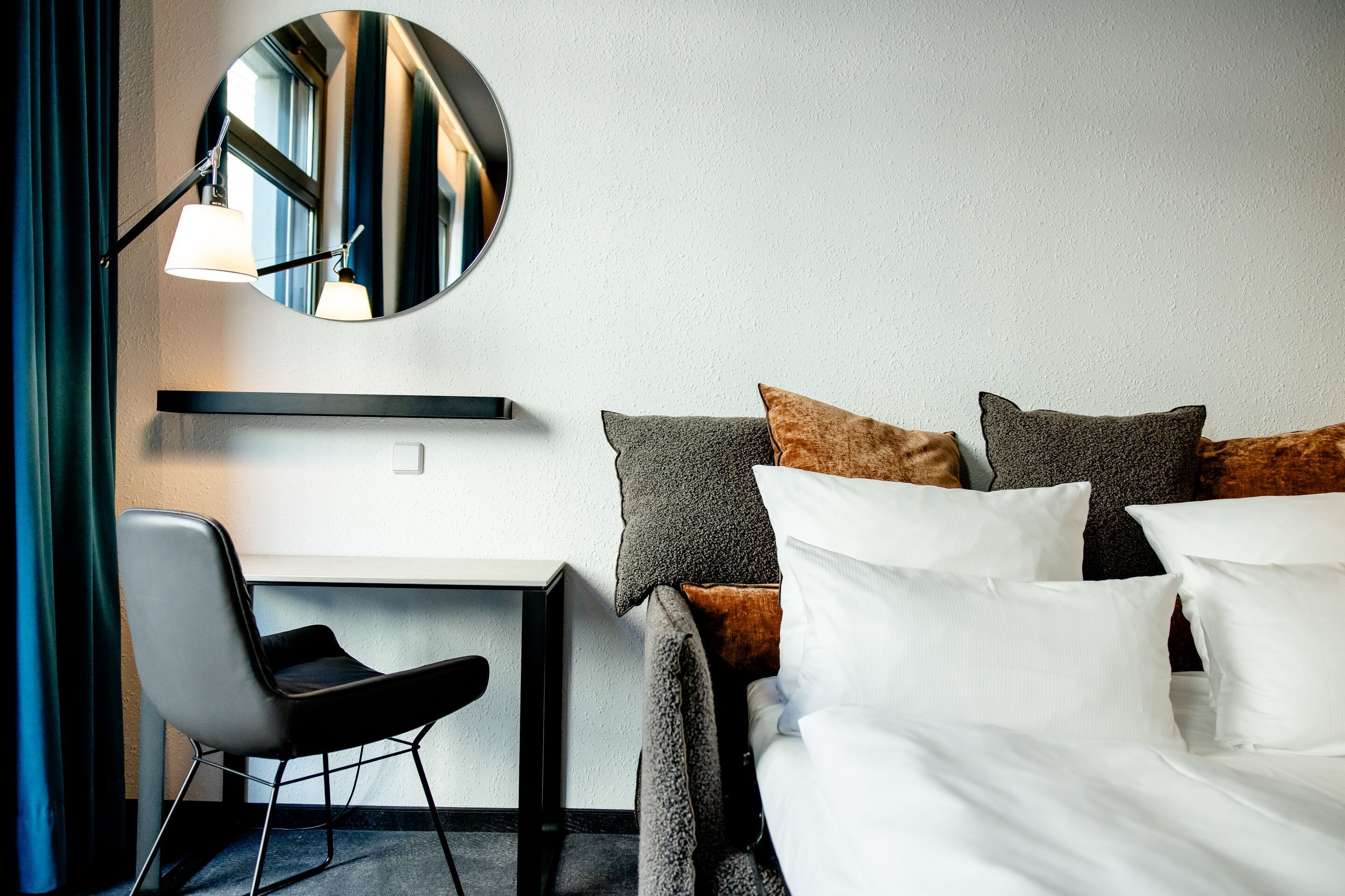 Hotel Leipzig Nikolaikirche Motel One Hotelzimmer mit gemütlichem Bett, mehrfarbigen Kissen, einem schwarzen Schreibtisch und Stuhl sowie einem runden Wandspiegel, der warmes Licht reflektiert und eine einladende Atmosphäre schafft.