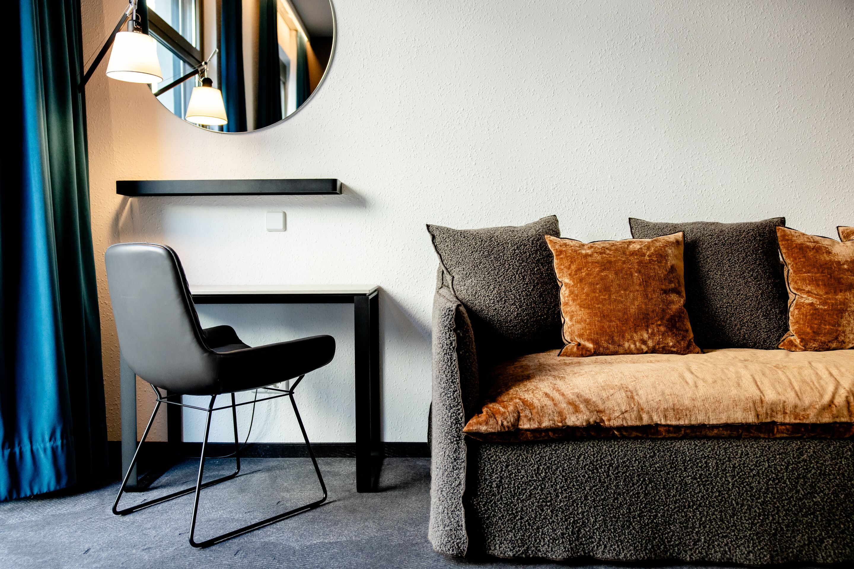 Hotel Leipzig Nikolaikirche Motel One gemütliche Sitzecke mit weichem, grauem Sofa und braunen Kissen, daneben ein moderner Stuhl vor kleinem schwarzen Tisch, runder Wandspiegel und sanfte Beleuchtung schaffen entspannte Atmosphäre.