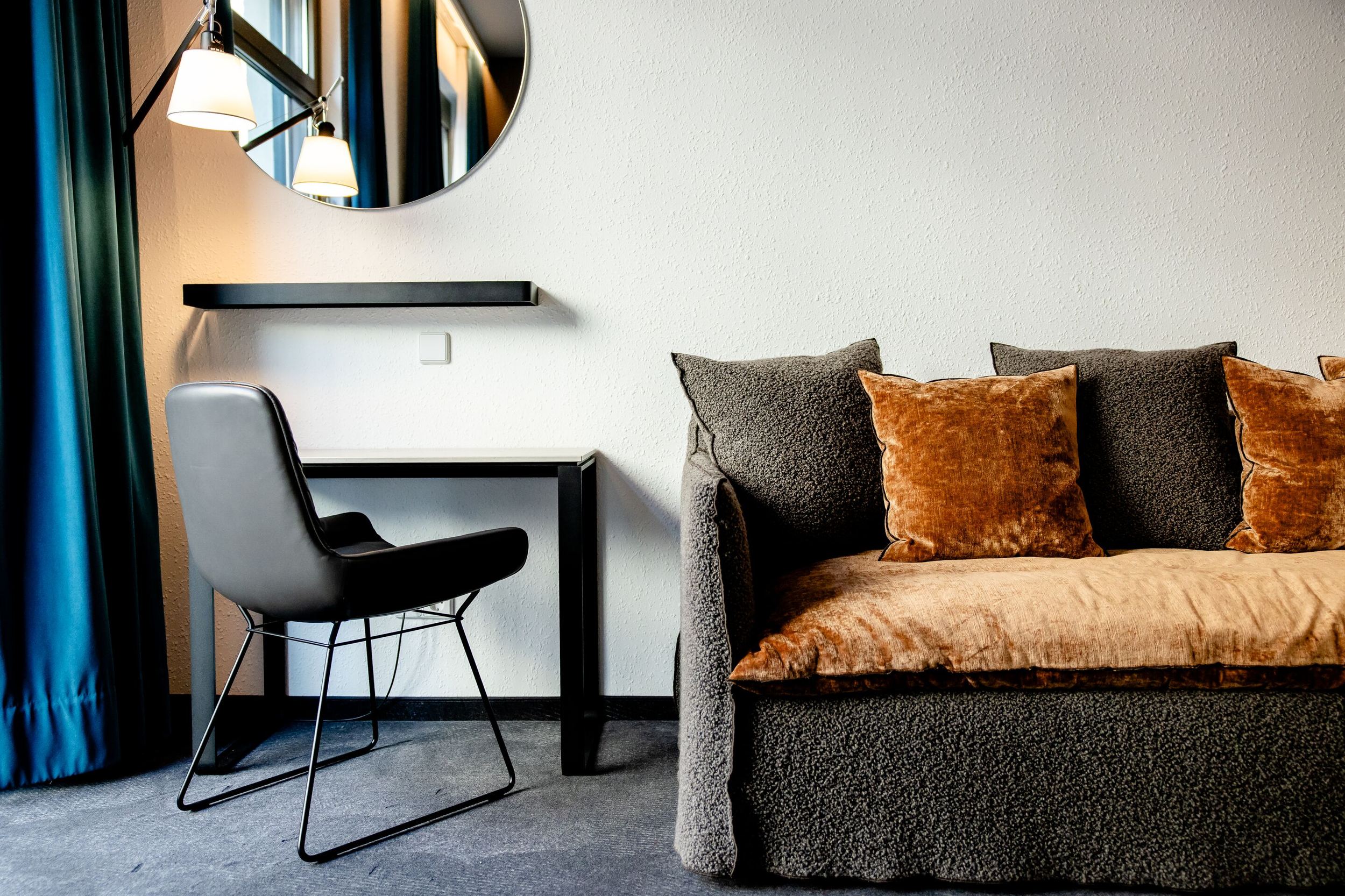 Hotel Leipzig Nikolaikirche Motel One gemütliche Sitzecke mit weichem, grauem Sofa und braunen Kissen, daneben ein moderner Stuhl vor kleinem schwarzen Tisch, runder Wandspiegel und sanfte Beleuchtung schaffen entspannte Atmosphäre.