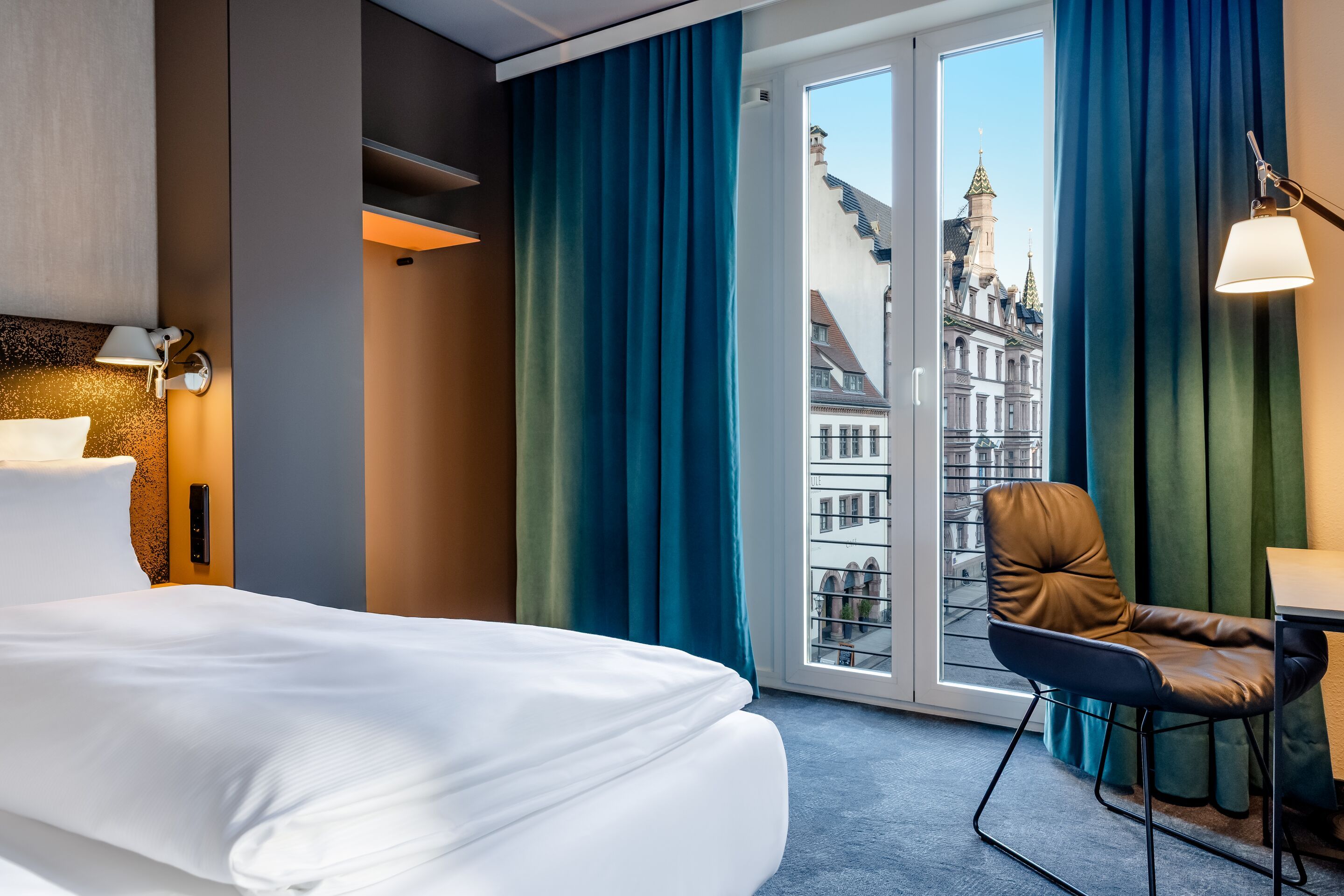 Hotel Leipzig Nikolaikirche Motel One Zimmer mit großem Bett, bequemen Sessel und bodentiefen Fenstern, die einen Blick auf historische Stadtarchitektur bieten. Helle, ruhige Atmosphäre mit gedeckten Farben und angenehmer Beleuchtung.