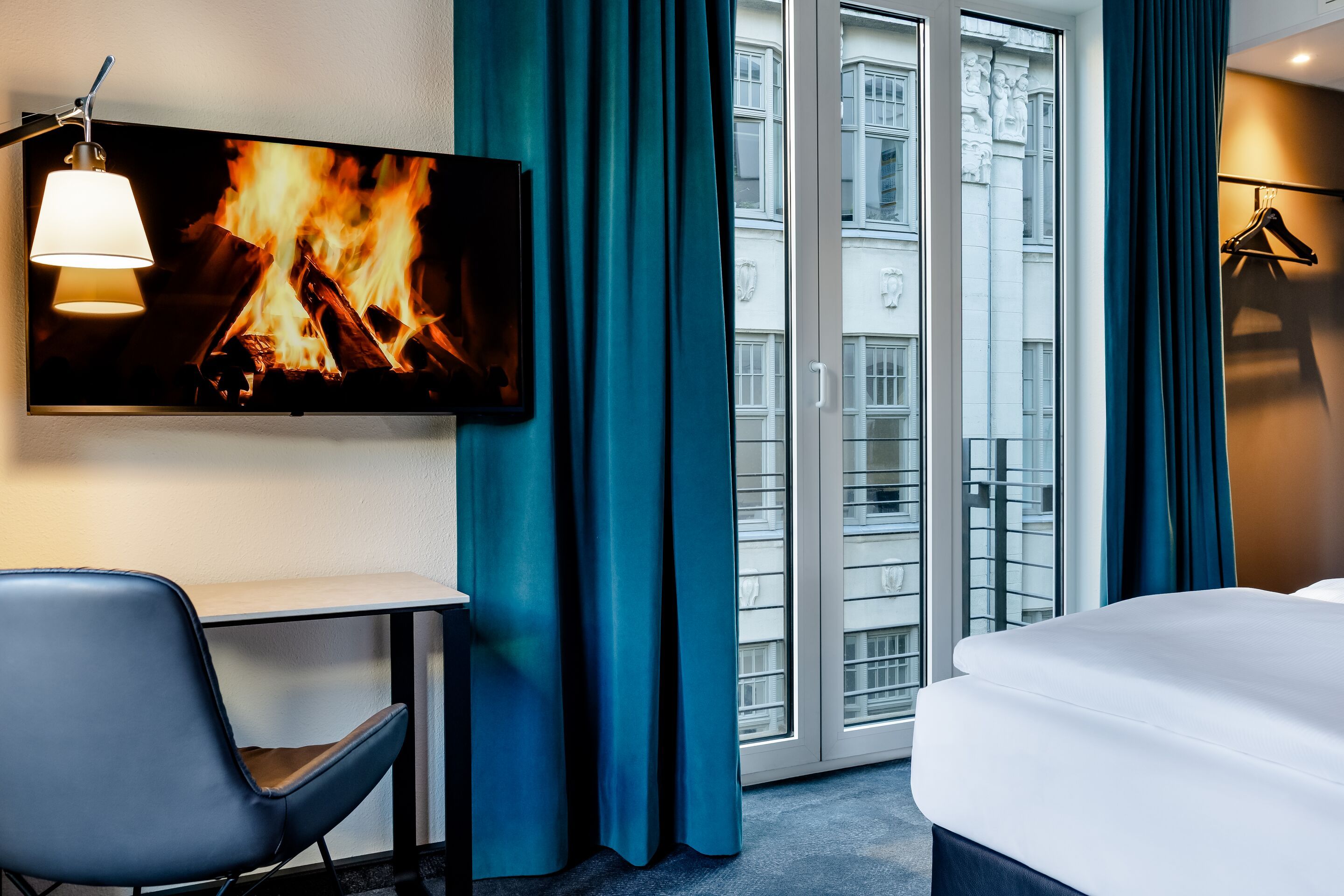 Hotel Leipzig Nikolaikirche Motel One helles, modernes Hotelzimmer mit gemütlichem Bett, großem Fenster zum Stadtblick und einem Fernseher, der ein Kaminfeuer zeigt, für entspannte Abende in stilvollem Ambiente.