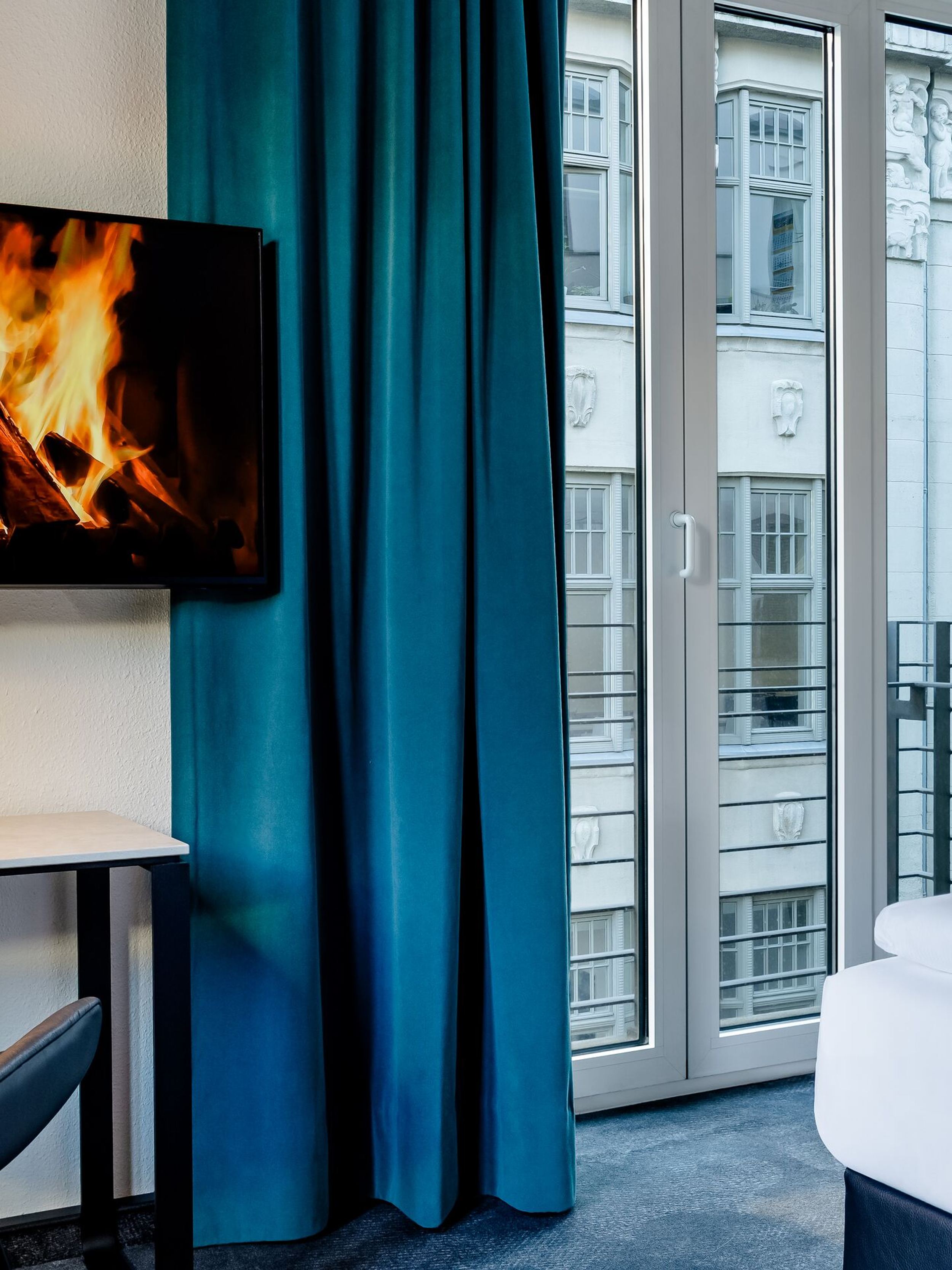 Hotel Leipzig Nikolaikirche Motel One helles, modernes Hotelzimmer mit gemütlichem Bett, großem Fenster zum Stadtblick und einem Fernseher, der ein Kaminfeuer zeigt, für entspannte Abende in stilvollem Ambiente.