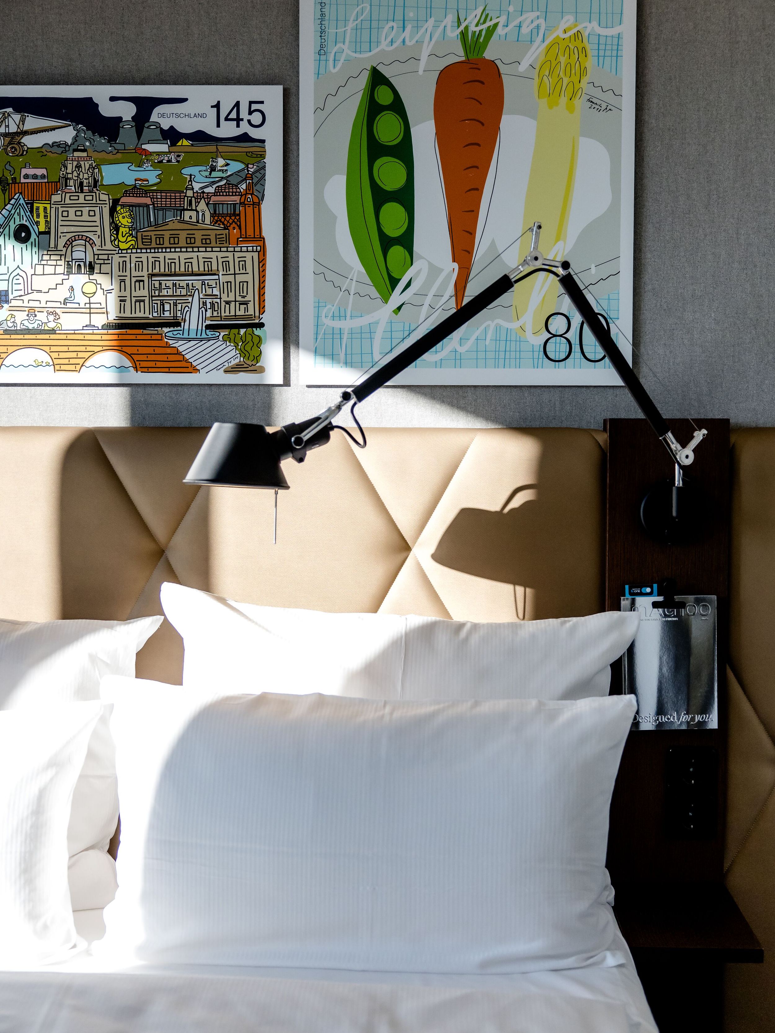 Hotel Leipzig Post Motel One Bett mit weißer Bettwäsche vor gepolstertem Kopfteil, darüber zwei bunte Kunstdrucke, eine mit Gemüsedarstellungen und eine mit Städtemotiv. An der Wand befestigte verstellbare Leselampe. Tageslicht fällt auf das Bett.