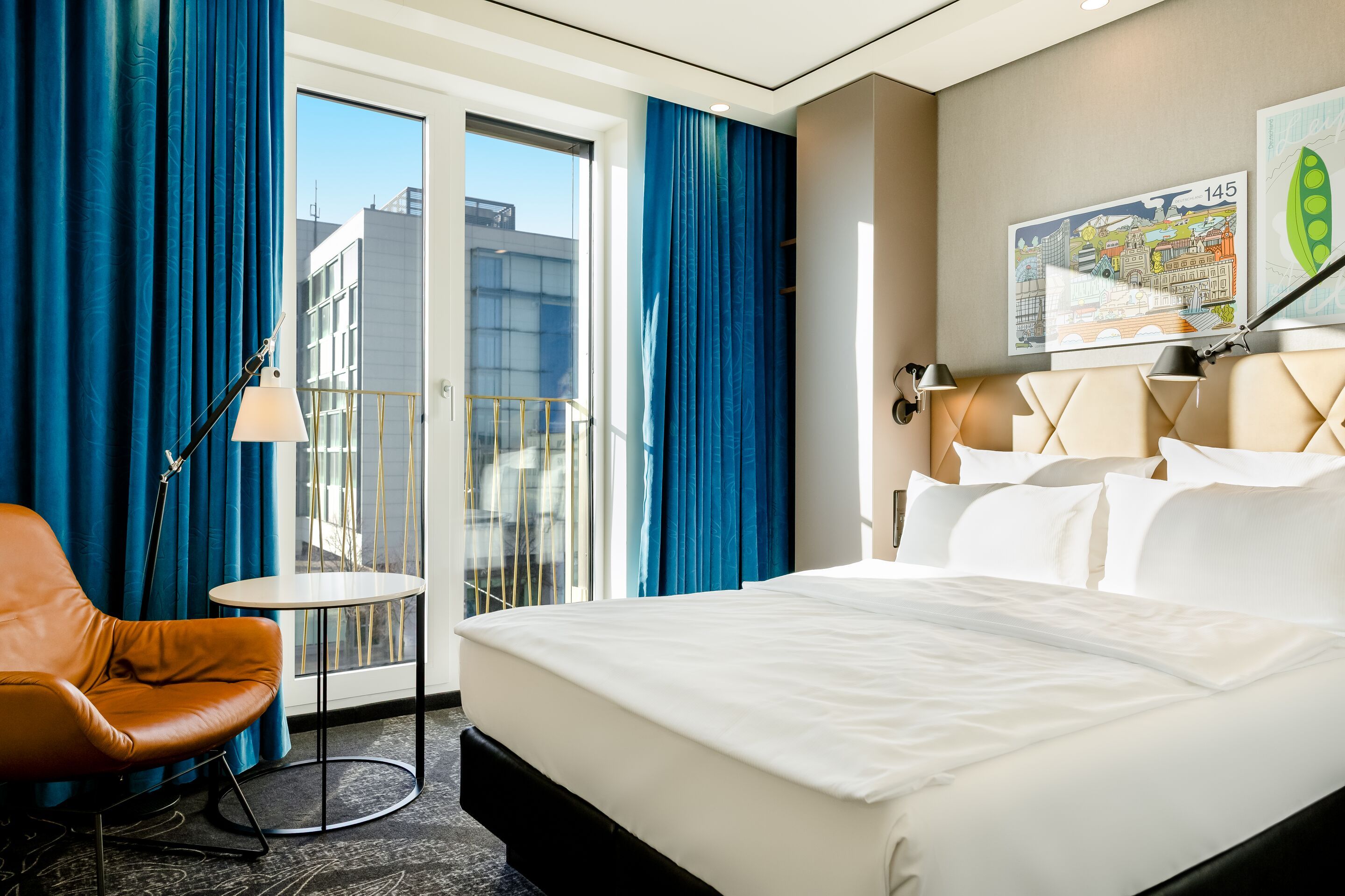 Hotel Leipzig Post Motel One modern room with a large bed, comfortable leather armchair and a small round table in front of floor-to-ceiling windows overlooking the city. Bright, friendly atmosphere thanks to natural lighting and stylish blue curtains.