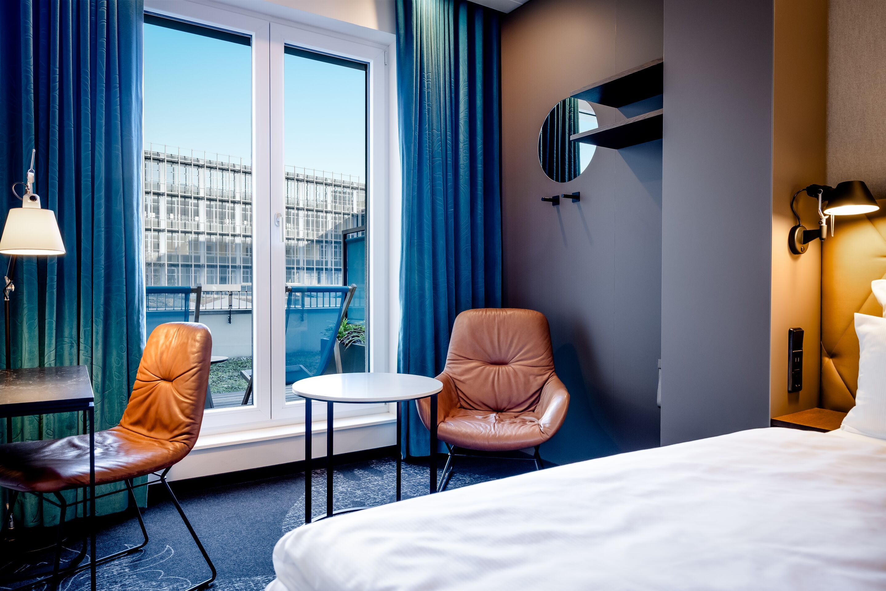 Hotel Leipzig Post Motel One modern hotel room with large window and view of the outdoors and balcony, two comfortable leather armchairs, small round table and a calming, bright atmosphere for relaxed working or relaxation.