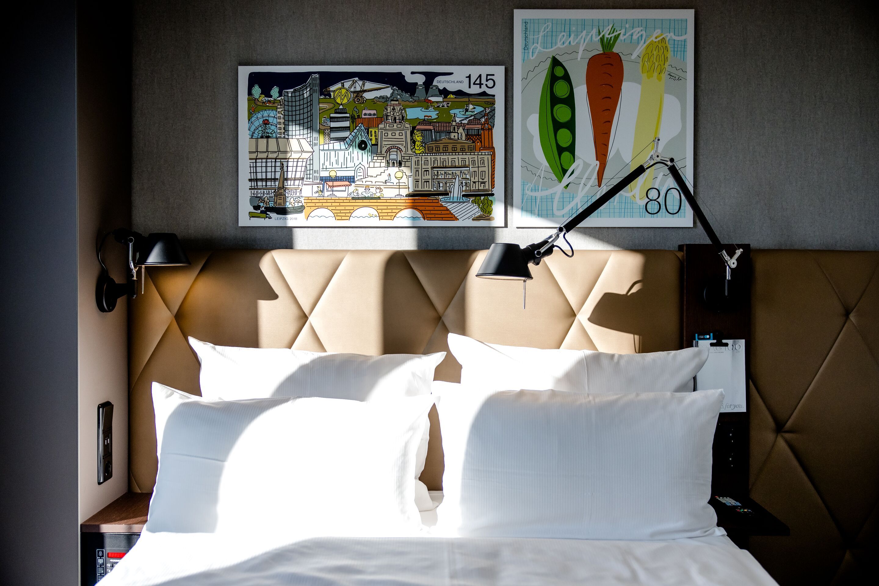 Hotel Motel One Leipzig-Post Hell beleuchtetes Doppelbett mit weißen Kissen und Bettwäsche, gepolstertem braunem Kopfteil, zwei verstellbaren Wandlampen und moderner Kunst mit farbenfrohen Motiven an der Wand.