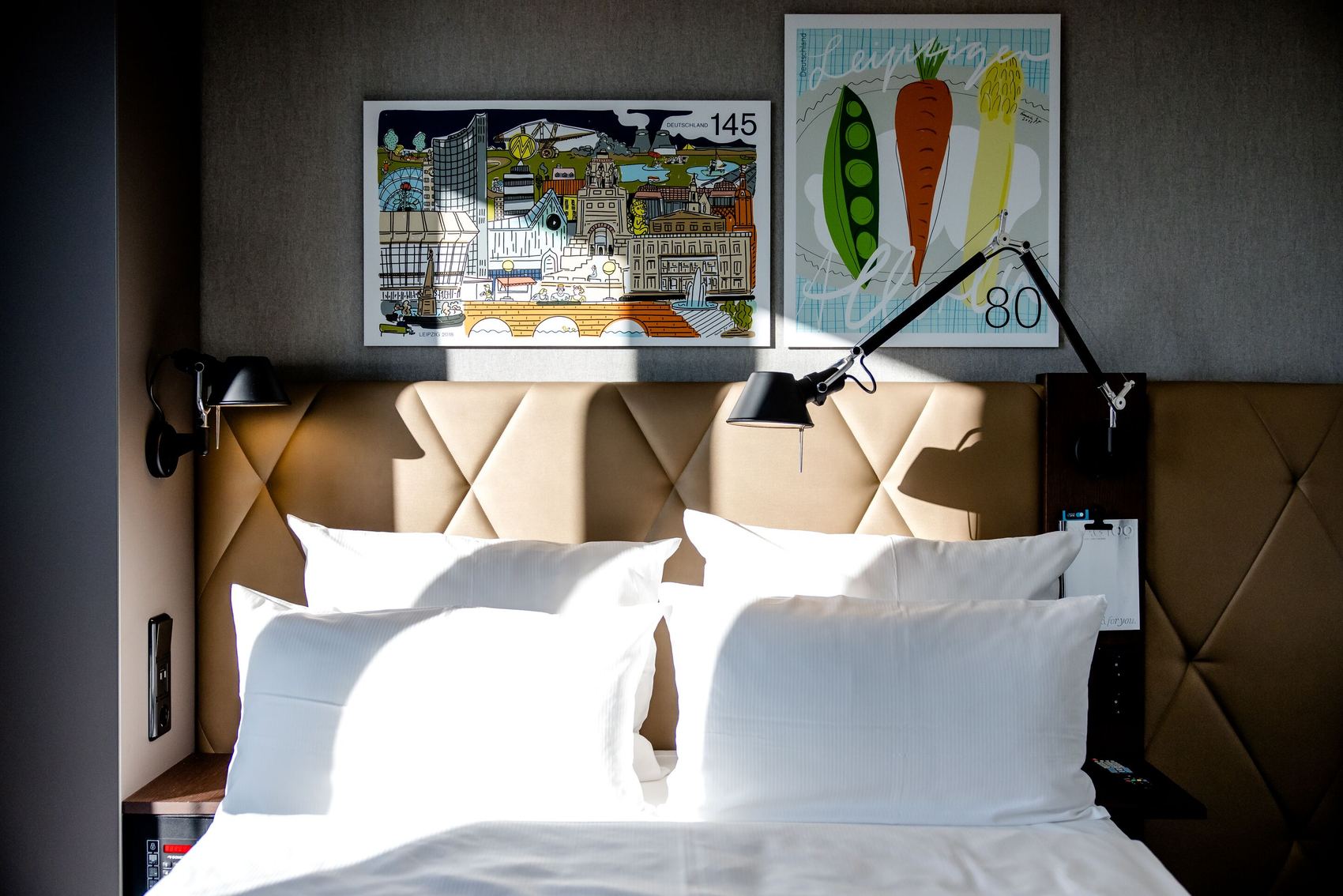 Hotel Motel One Leipzig-Post Hell beleuchtetes Doppelbett mit weißen Kissen und Bettwäsche, gepolstertem braunem Kopfteil, zwei verstellbaren Wandlampen und moderner Kunst mit farbenfrohen Motiven an der Wand.