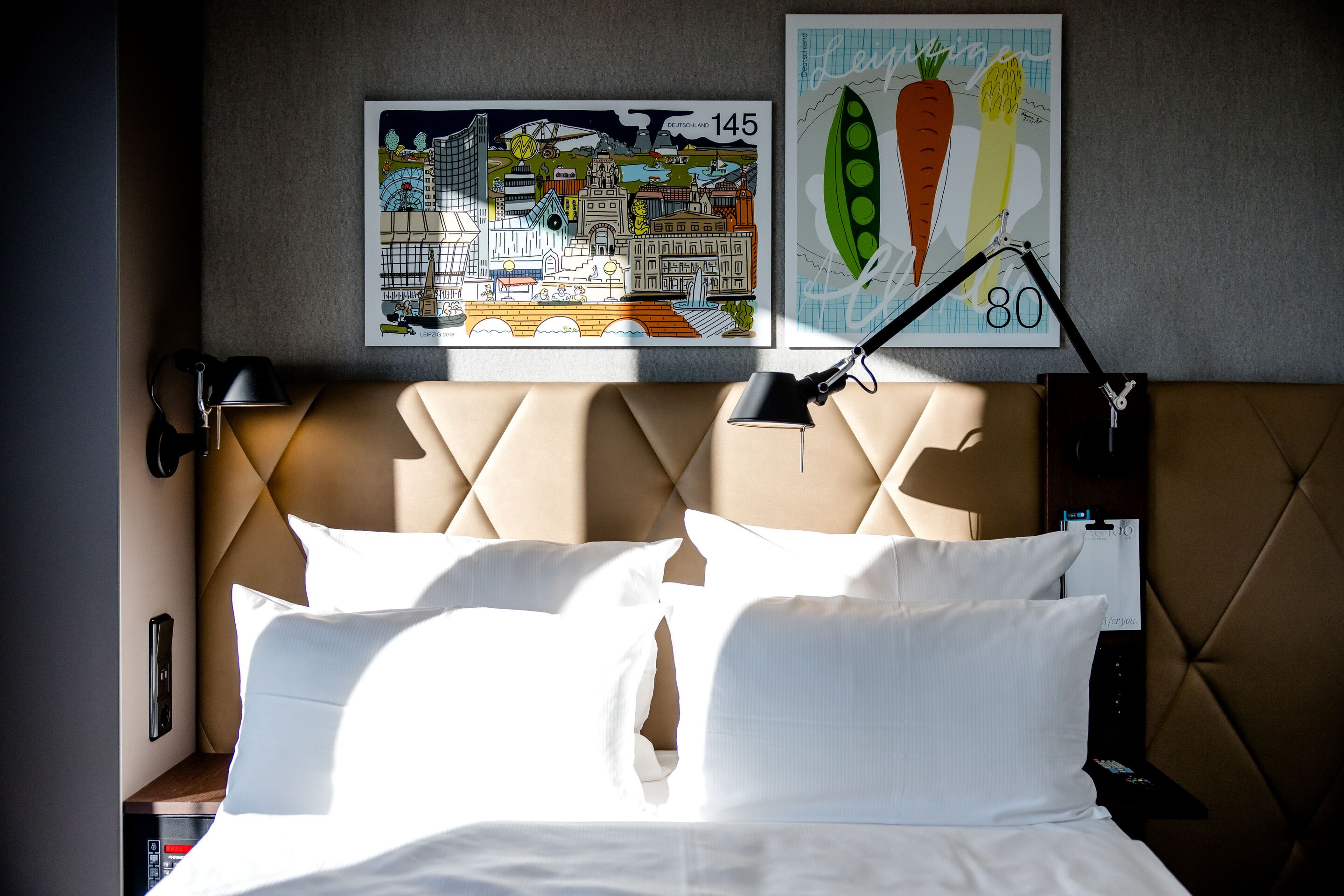 Hotel Motel One Leipzig-Post Hell beleuchtetes Doppelbett mit weißen Kissen und Bettwäsche, gepolstertem braunem Kopfteil, zwei verstellbaren Wandlampen und moderner Kunst mit farbenfrohen Motiven an der Wand. Hotel Motel One Leipzig-Post Hell beleuchtetes Doppelbett mit weißen Kissen und Bettwäsche, gepolstertem braunem Kopfteil, zwei verstellbaren Wandlampen und moderner Kunst mit farbenfrohen Motiven an der Wand.