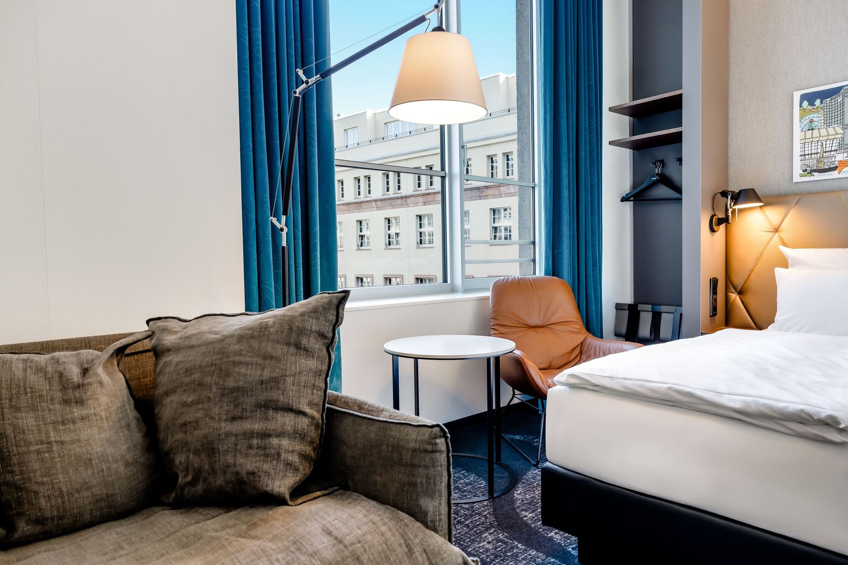 Hotel Motel One Leipzig-Post Helles Hotelzimmer mit gemütlichem Sofa, Lederstuhl, rundem Tisch, großem Fenster mit Blick auf Stadtgebäude und einem komfortablen Bett mit weißen Bettwäsche und Leselampe.