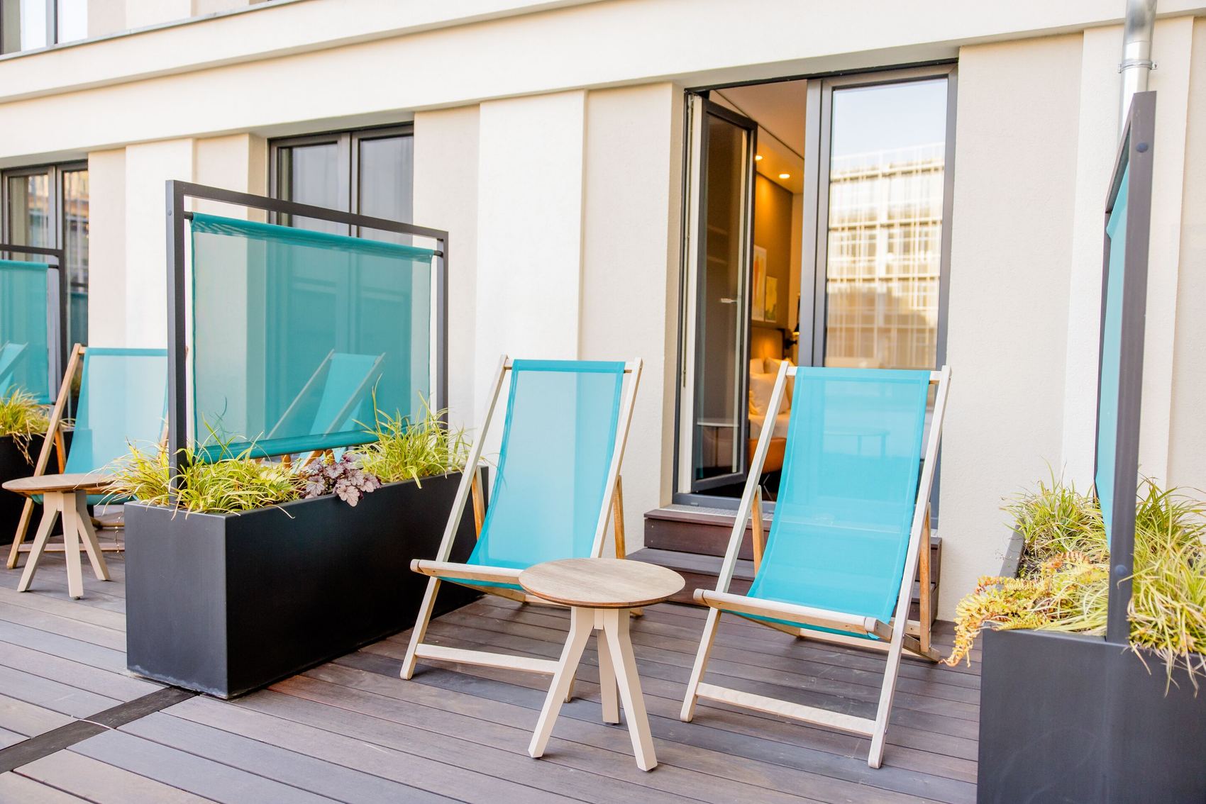 Hotel Motel One Leipzig-Post Terrassenbereich mit zwei bequemen Liegestühlen und kleinem Tisch, umgeben von modernen Pflanzgefäßen, ideal zum Entspannen mit Blick ins Freie. Zugang zu einem hellen Hotelzimmer durch eine geöffnete Tür.