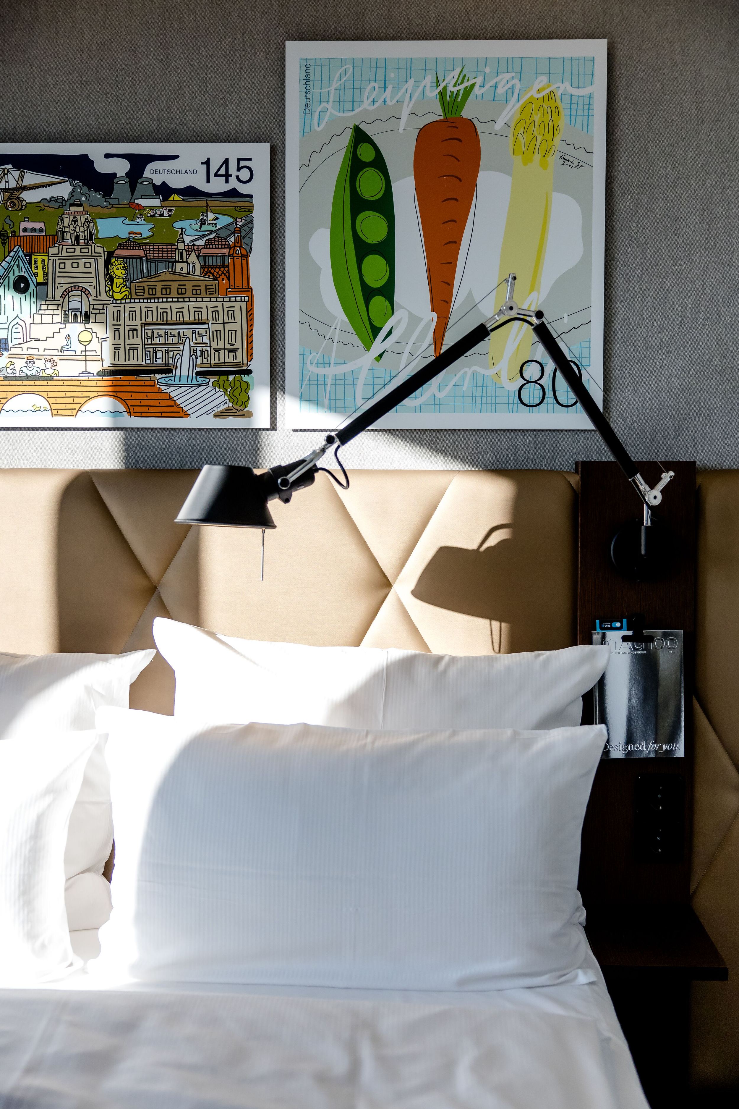 Hotel Leipzig Post Motel One Bett mit weißer Bettwäsche vor gepolstertem Kopfteil, darüber zwei bunte Kunstdrucke, eine mit Gemüsedarstellungen und eine mit Städtemotiv. An der Wand befestigte verstellbare Leselampe. Tageslicht fällt auf das Bett.