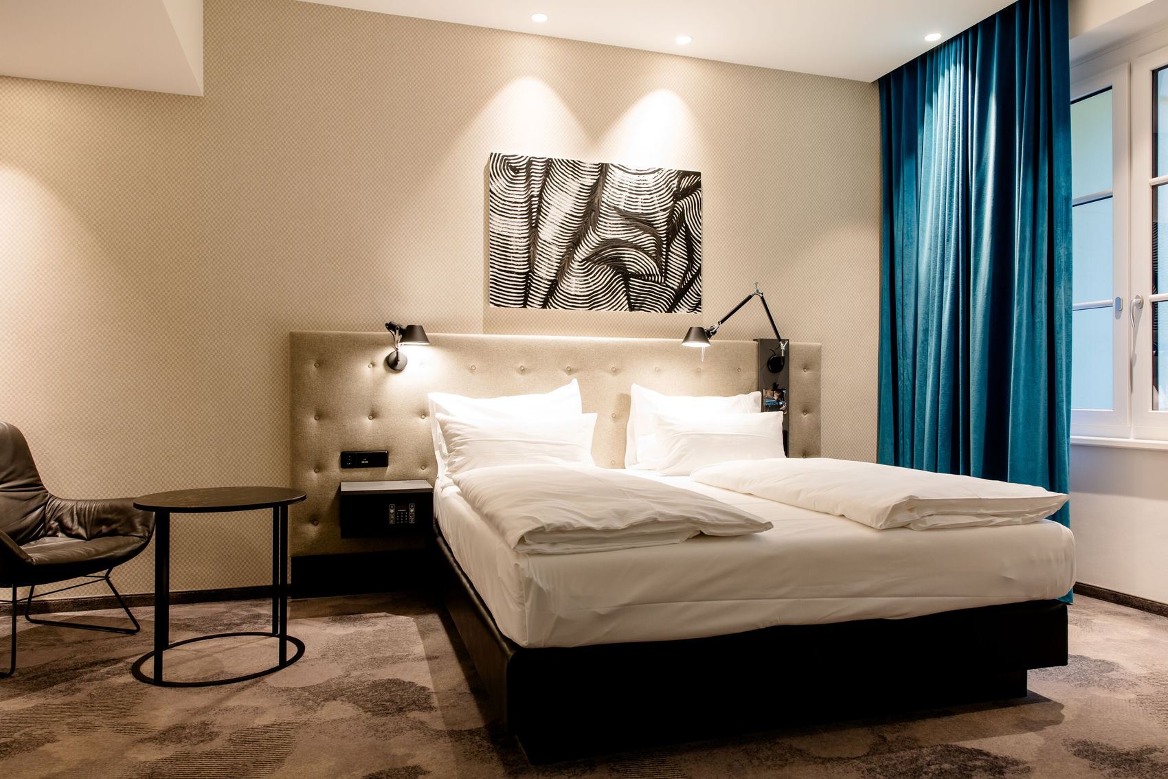 Hotel Motel One Linz-Hauptplatz Hell gestaltetes Hotelzimmer mit zwei Einzelbetten, modernem Kopfteil und stilvoller Beleuchtung, daneben ein Sessel und ein Beistelltisch, großer Fensterbereich mit blauen Vorhängen für angenehme Tageslichtgestaltung.
