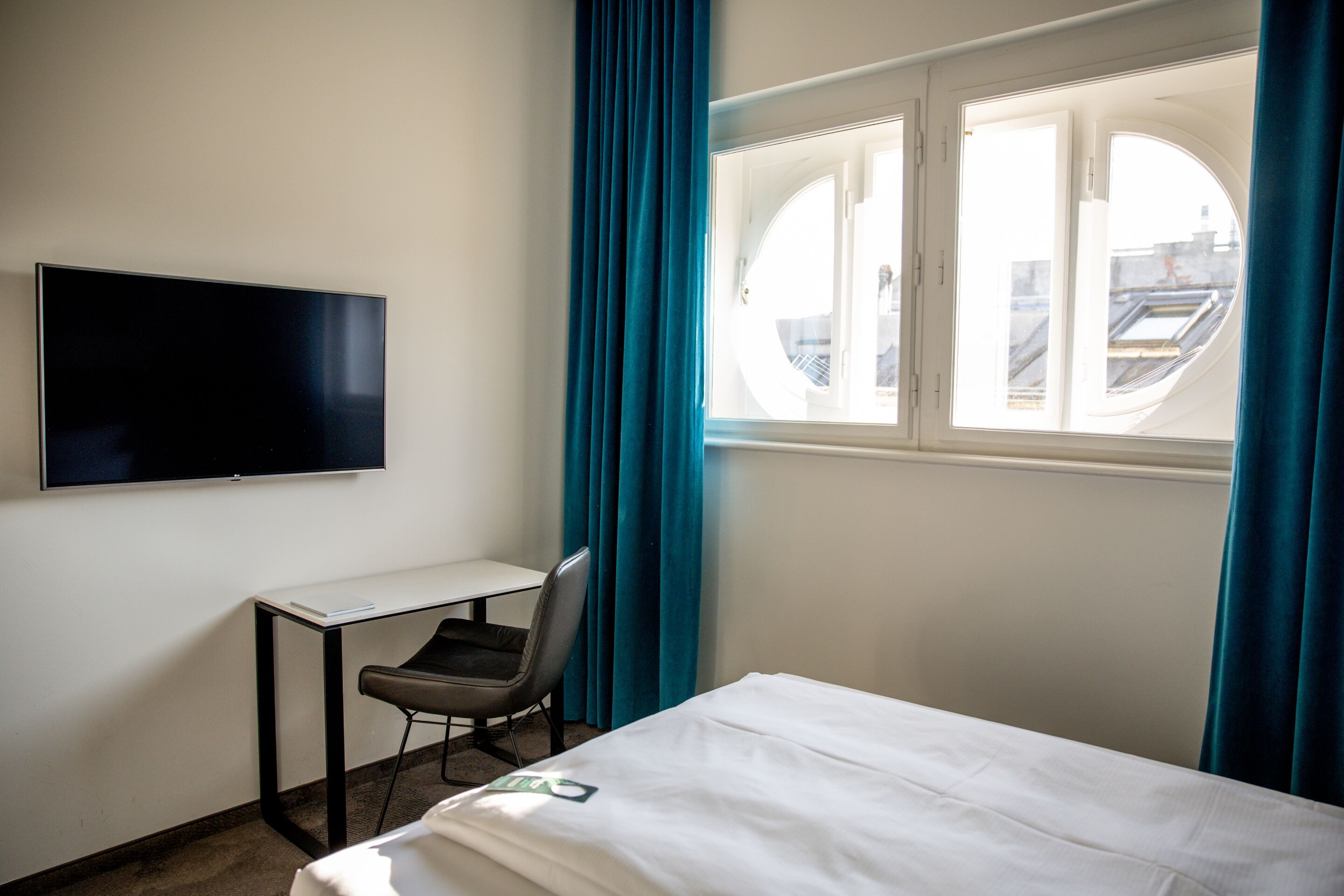 Helles Hotelzimmer mit gemütlichem Bett, großem Fenster mit Blick auf das Stadtpanorama, Arbeitsplatz mit Stuhl und wandmontiertem Fernseher, ergänzt durch dunkle Vorhänge für angenehme Ruhe und Privatsphäre.