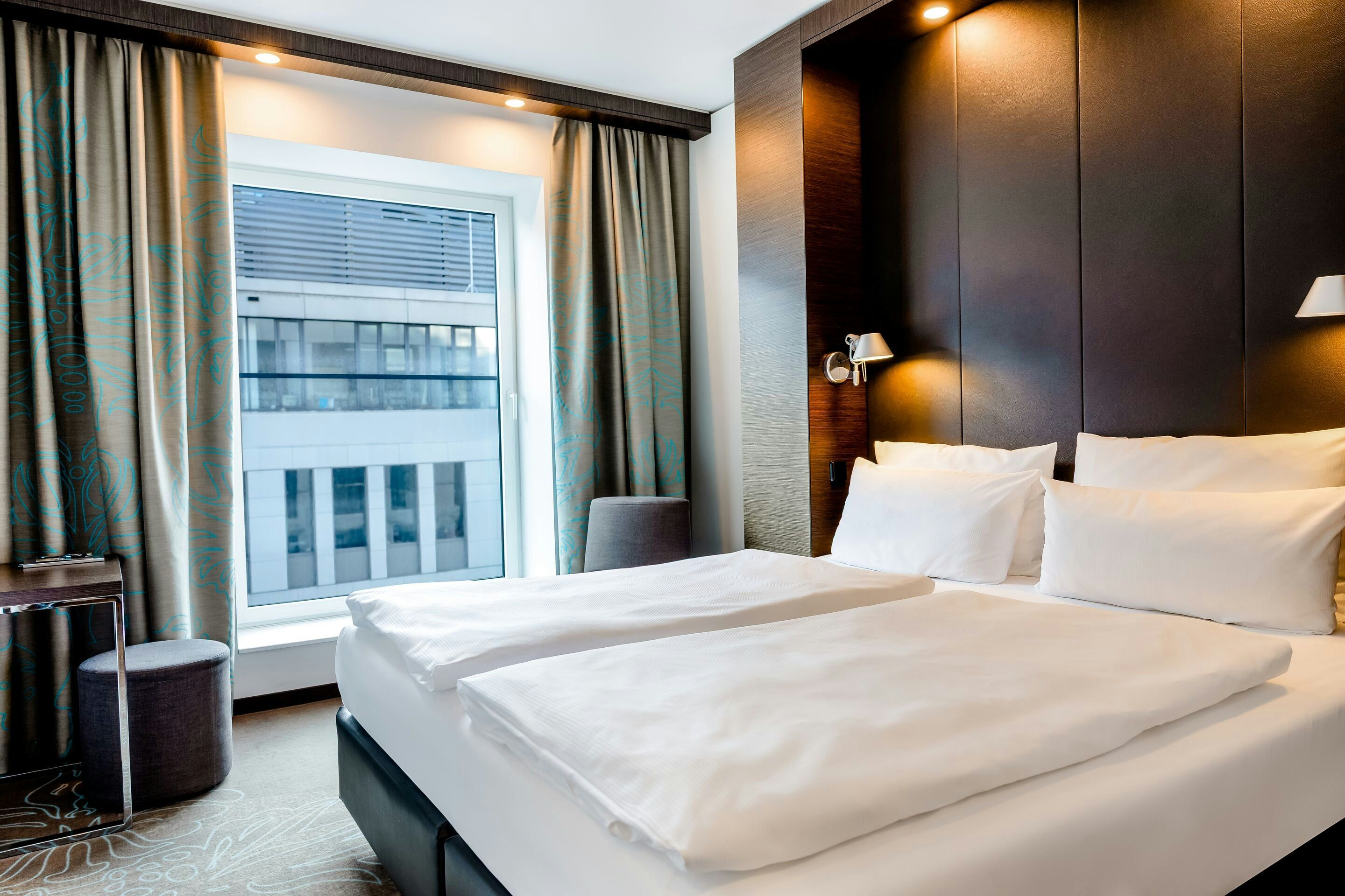 Modernes Hotelzimmer mit großem, bequemem Doppelbett, weichen Kopfkissen und sanfter Beleuchtung. Großes Fenster mit Blick auf städtische Gebäude, natürliche Lichtquelle. Ruhige, elegante Atmosphäre für erholsamen Aufenthalt.