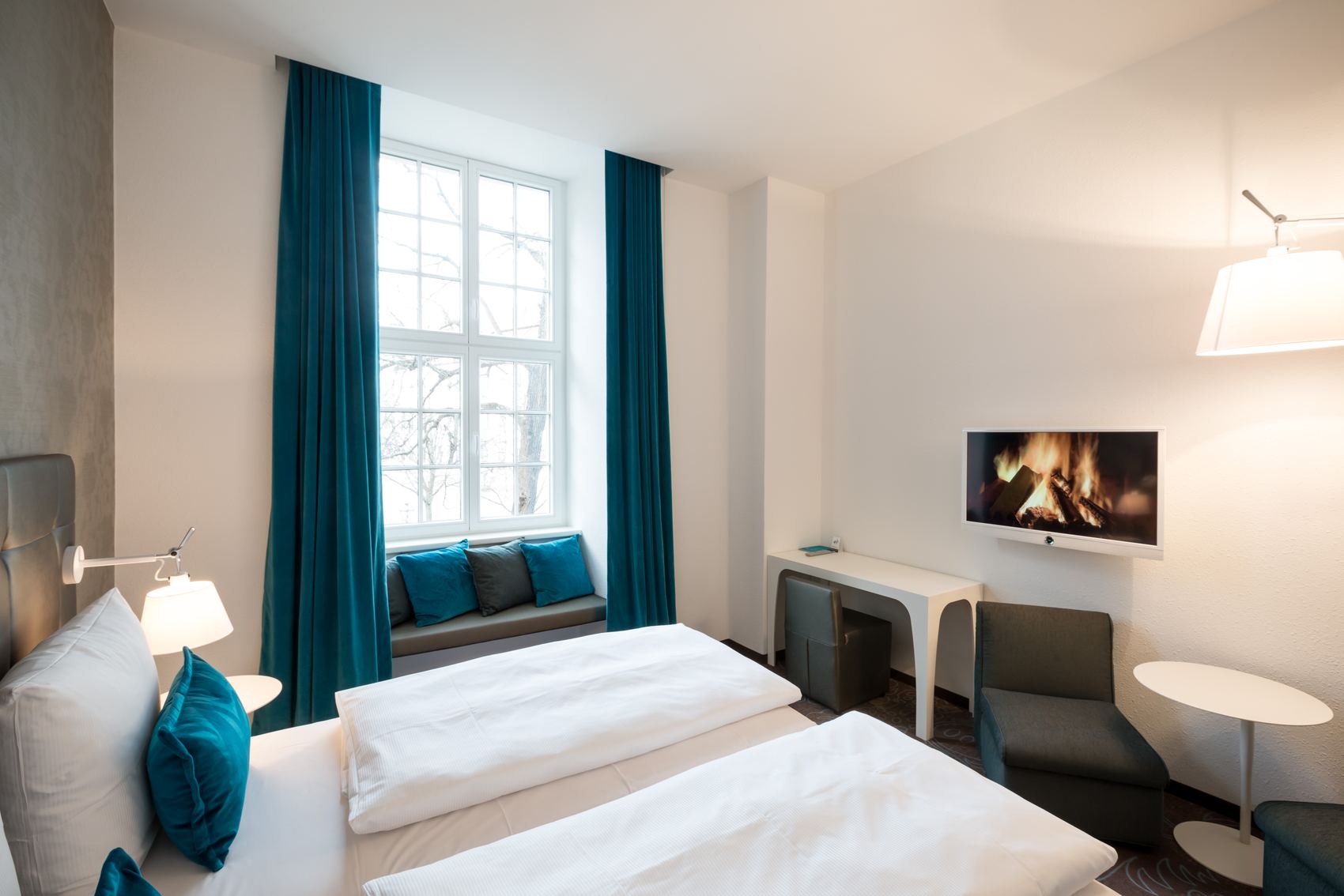 Hotel Motel One Magdeburg Helles Hotelzimmer mit zwei Einzelbetten, gemütlicher Fensterbank mit Kissen, blauem Vorhang, Sitzecke und Fernseher, der ein Feuer zeigt. Ruhige, einladende Atmosphäre für Entspannung und Erholung.