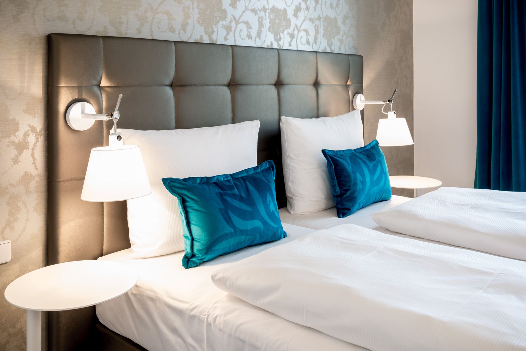 Hotel Motel One Magdeburg Zwei gemütliche Einzelbetten mit weißen Bettlaken, je zwei weißen und blauen Kissen, Kopfteil in Lederoptik und je einer modernen Wandlampe, die eine warme, einladende Atmosphäre im Hotelzimmer schafft.