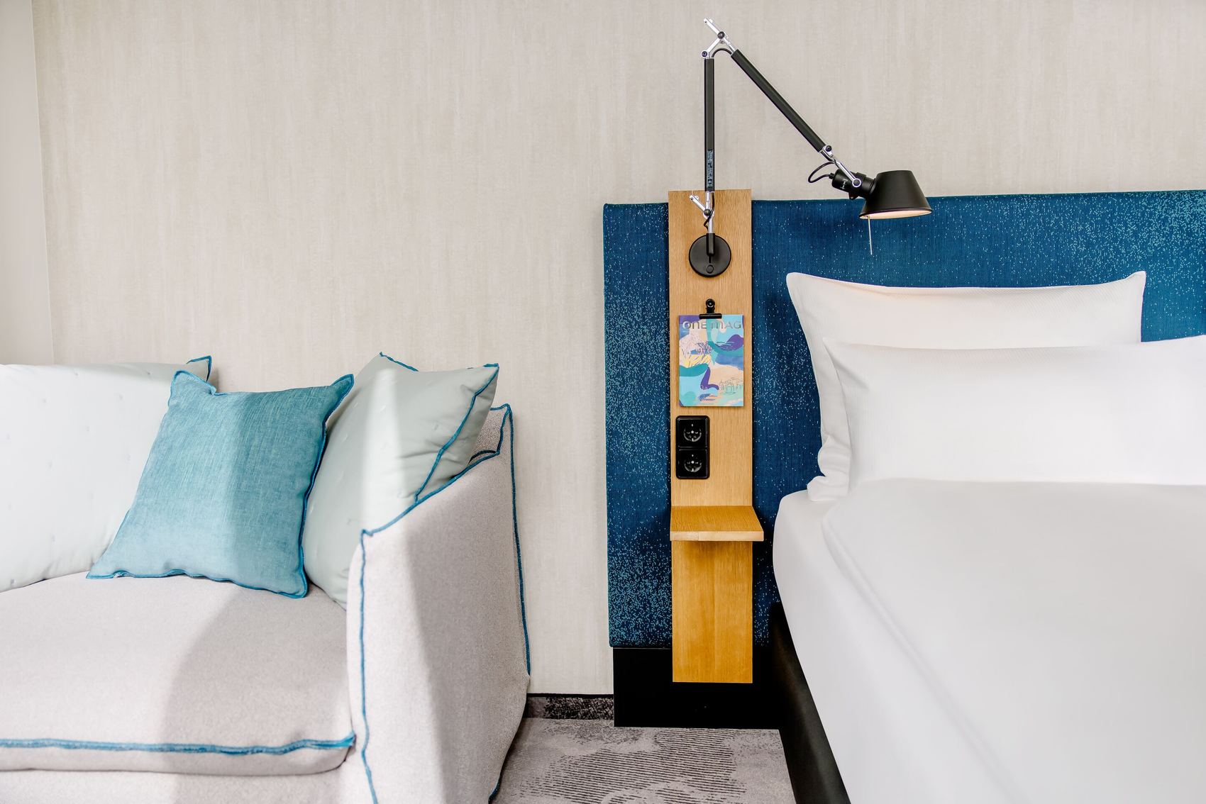 Hotel Motel One München-Campus Hell eingerichtetes Hotelzimmer mit gemütlichem Bett, blauer Stoffkopflehne, moderner Wandlampe und Sofa mit türkisfarbenen Kissen für entspannten Komfort.
