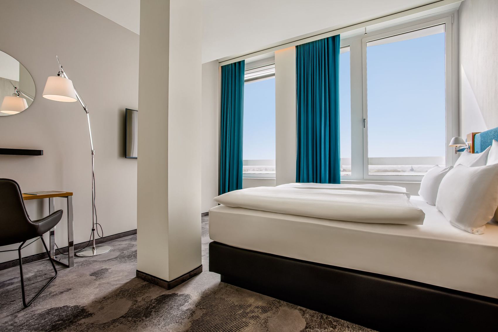 Hotel Motel One München-Campus Hell eingerichtetes Hotelzimmer mit großem Fenster, Blick aufs Meer, gemütlichem Doppelbett, modernen Möbeln und blauen Vorhängen für angenehme Atmosphäre und Entspannung.