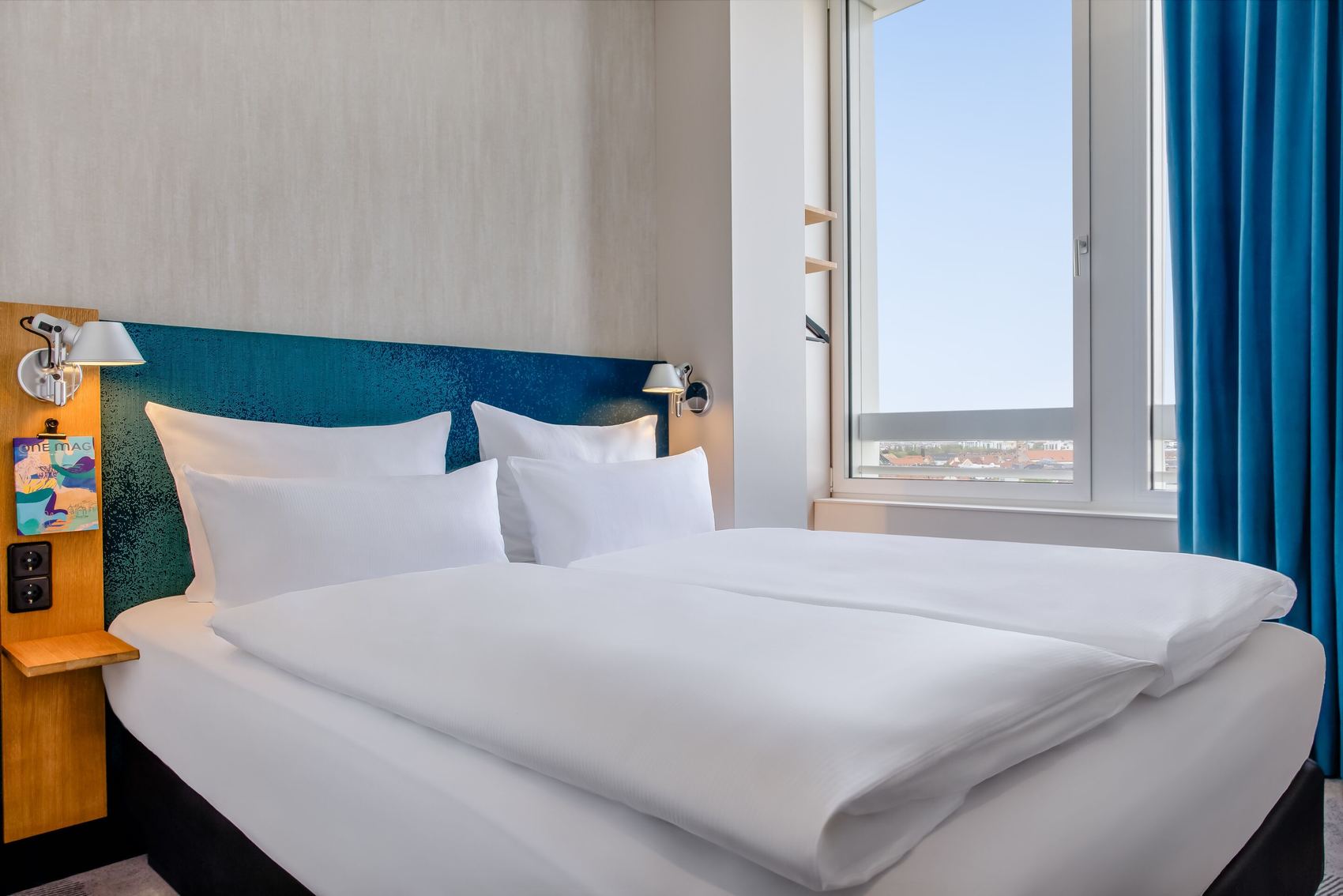 Hotel Motel One München-Campus Helles Hotelzimmer mit großem, bequemem Doppelbett, weißen Bettwäsche, modernem blauen Kopfteil und Fenster mit Tageslicht und Ausblick.