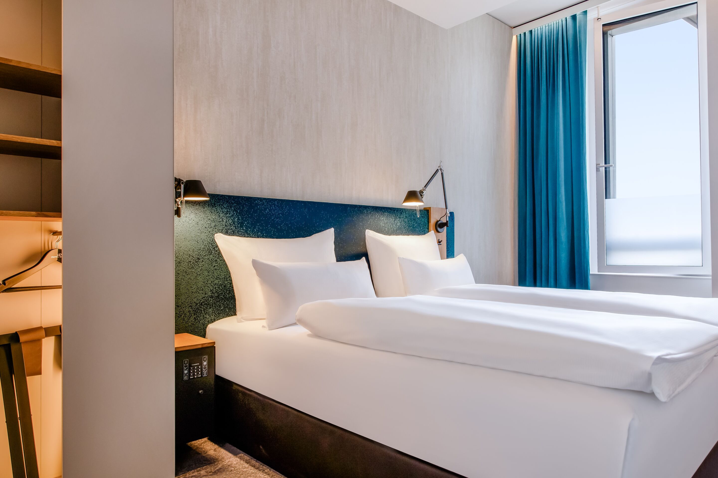 Hotel Motel One München-Campus Modern eingerichtetes Hotelzimmer mit großem, bequemem Doppelbett, hellem Ambiente und offenem Fenster für frische Luft.