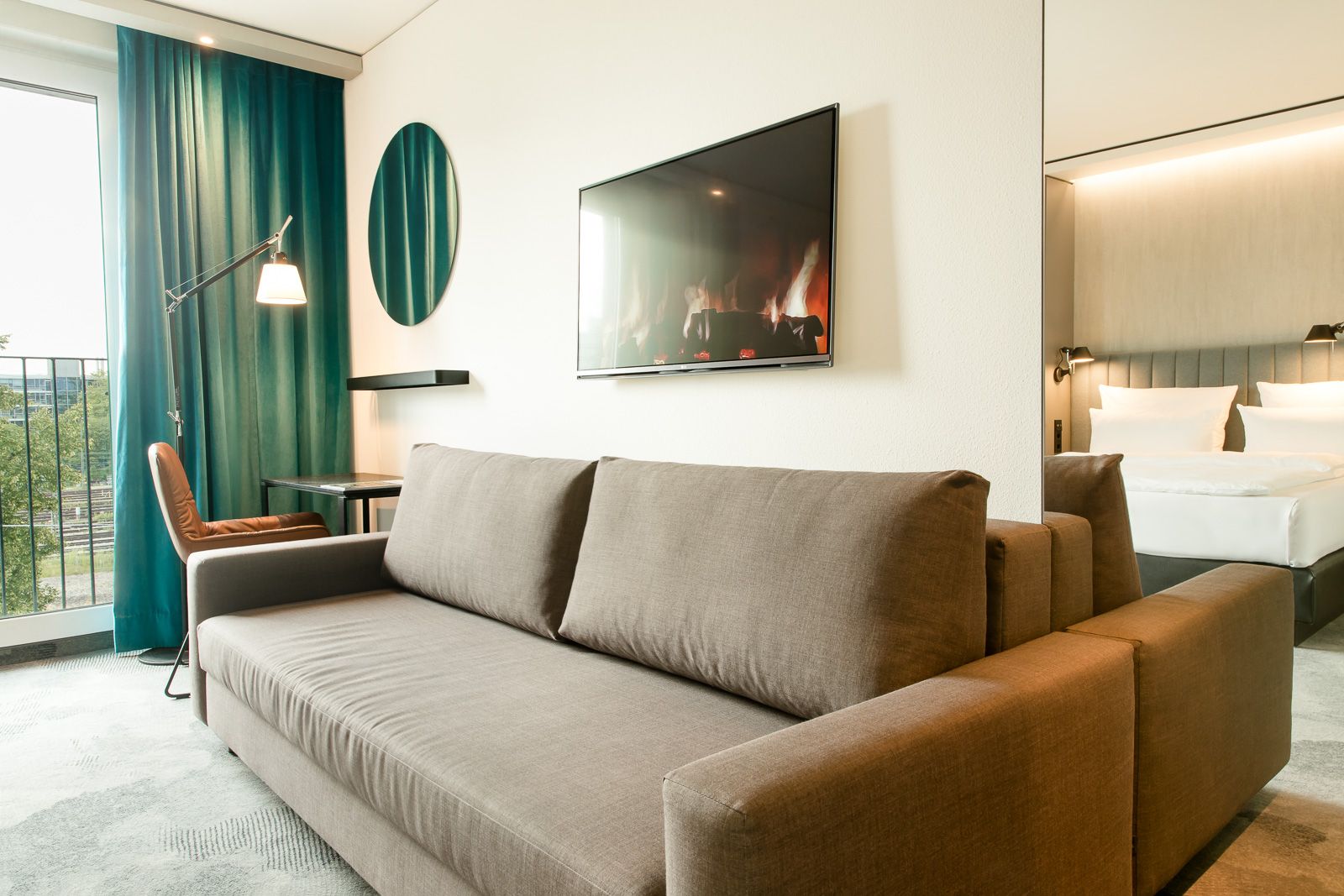 Designhotel München East Side Motel One großes und helles Zimmer mit Blick auf eine hellbraune Couch sowie Schreibtisch mit Stuhl