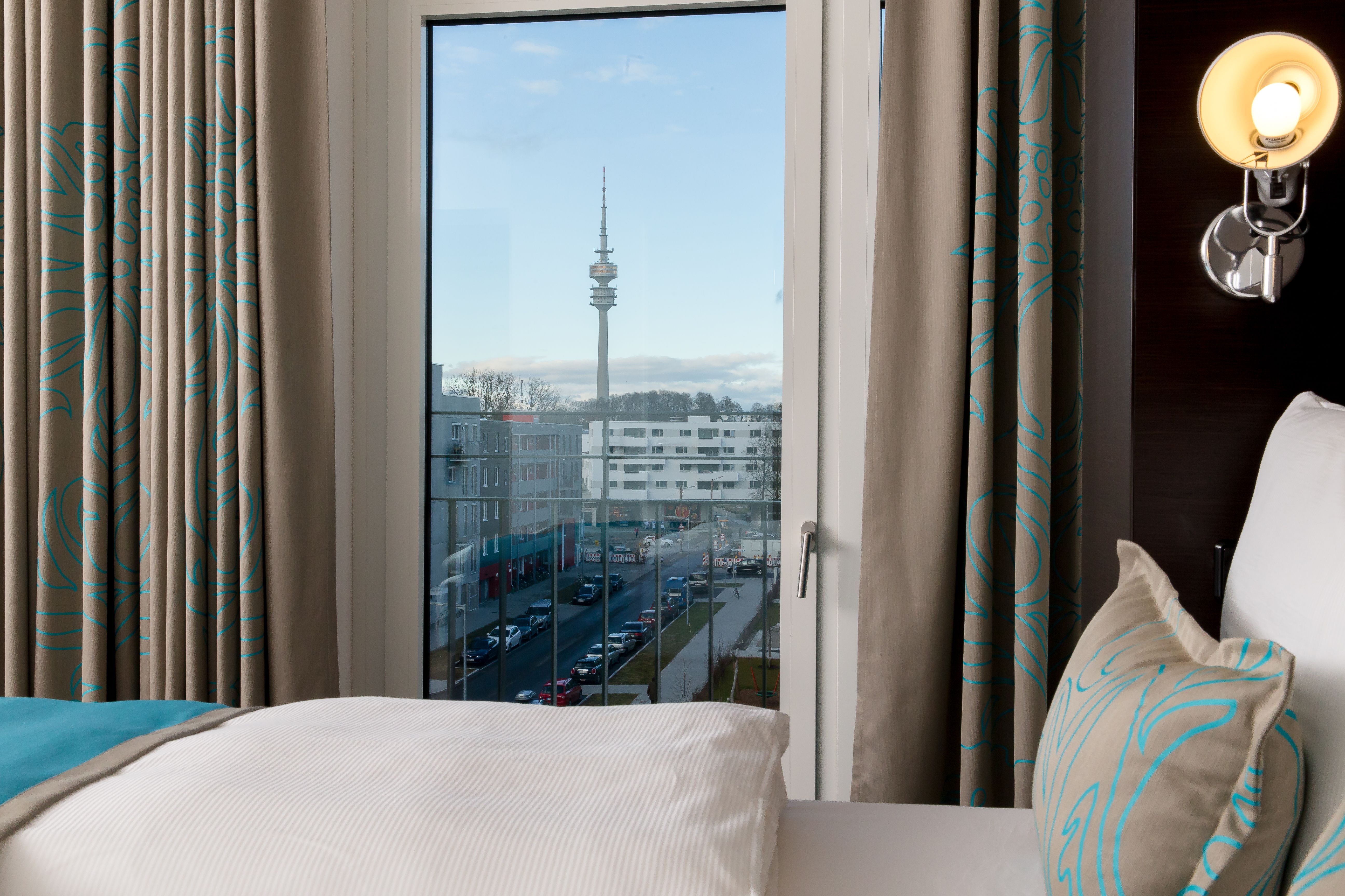 Hotel München Olympia Gate Motel One Zimmer mit Blick durch bodentiefes Fenster auf Olympiaturm