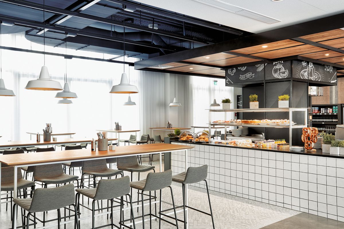 Modernes, helles Café mit hohen Thekentischen, gemütlichen Stühlen und einem Buffetbereich mit frischen Backwaren, Brezeln und Getränken. Angenehme Atmosphäre für Frühstück oder kleine Pausen.