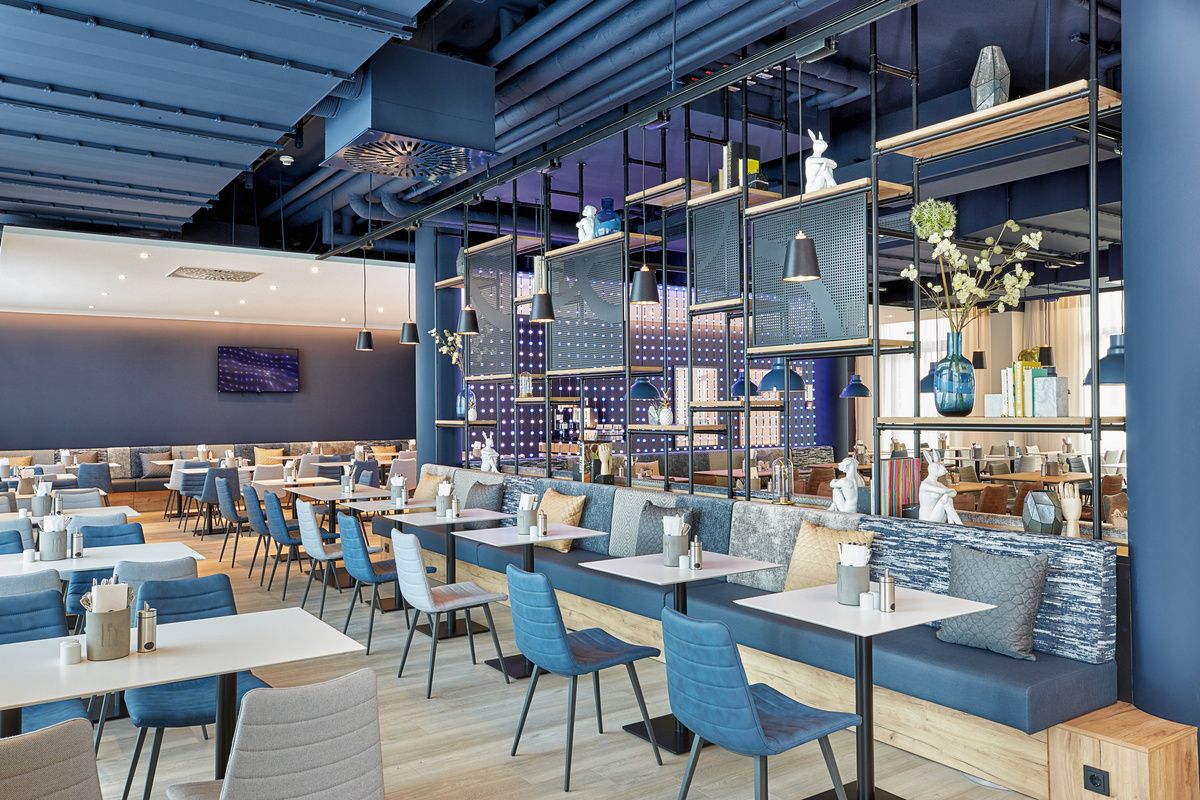 Modern eingerichteter Restaurantbereich mit hellen Tischen, blauen und grauen Stühlen sowie dekorativen Elementen auf Regalen und einer gemütlichen Atmosphäre.
