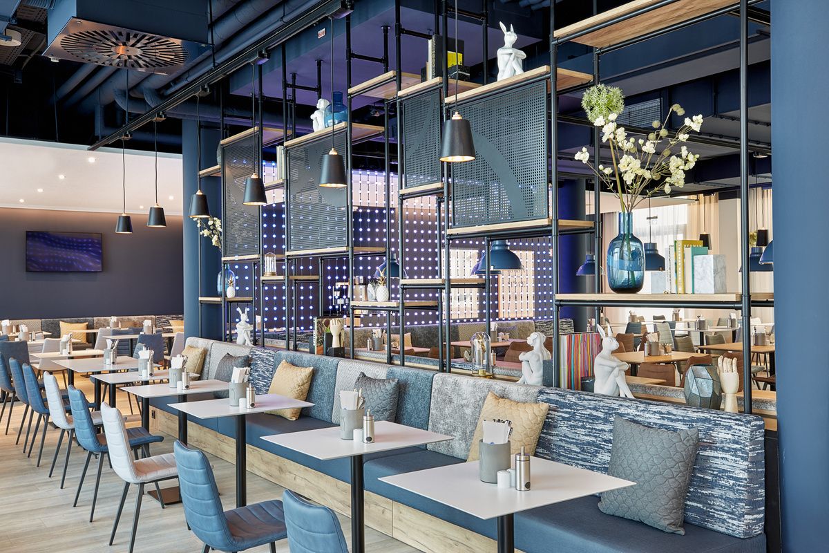 Modern eingerichteter Hotelrestaurantbereich mit bequemen Sitzbänken, blauen Stühlen, eleganter Beleuchtung und dekorativen Elementen, die eine ruhige und einladende Atmosphäre für Gäste schaffen.