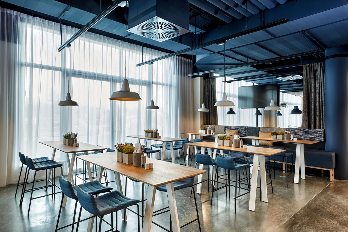 Helles, modernes Restaurant mit großen Fenstern, Holz- und Metalltischen, bequemen Stühlen und stilvollen Hängeleuchten. Angenehme Atmosphäre für entspannte Mahlzeiten oder Geschäftstreffen.