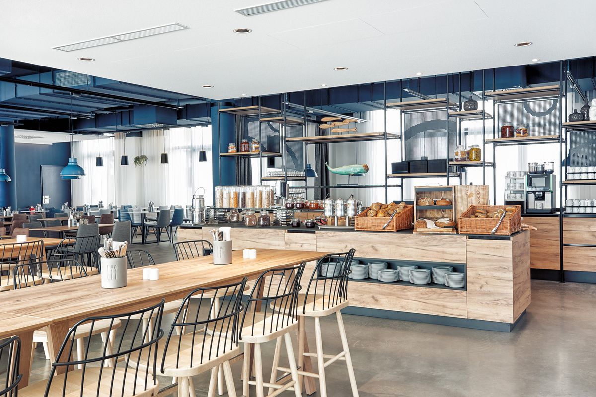 Heller, moderner Frühstücksbereich mit großen Holztischen, Metallstühlen und Buffets mit frischem Gebäck, Müsli und Kaffeemaschinen, der eine einladende Atmosphäre für Gäste schafft.