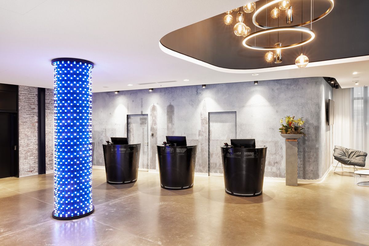 Moderner Hotellobby-Empfang mit drei schwarzen runden Rezeptionstheken, hellem Boden, einer dekorativen blauen Säule mit Lichtern und einer stilvollen Deckenbeleuchtung mit hängenden Kugeln.