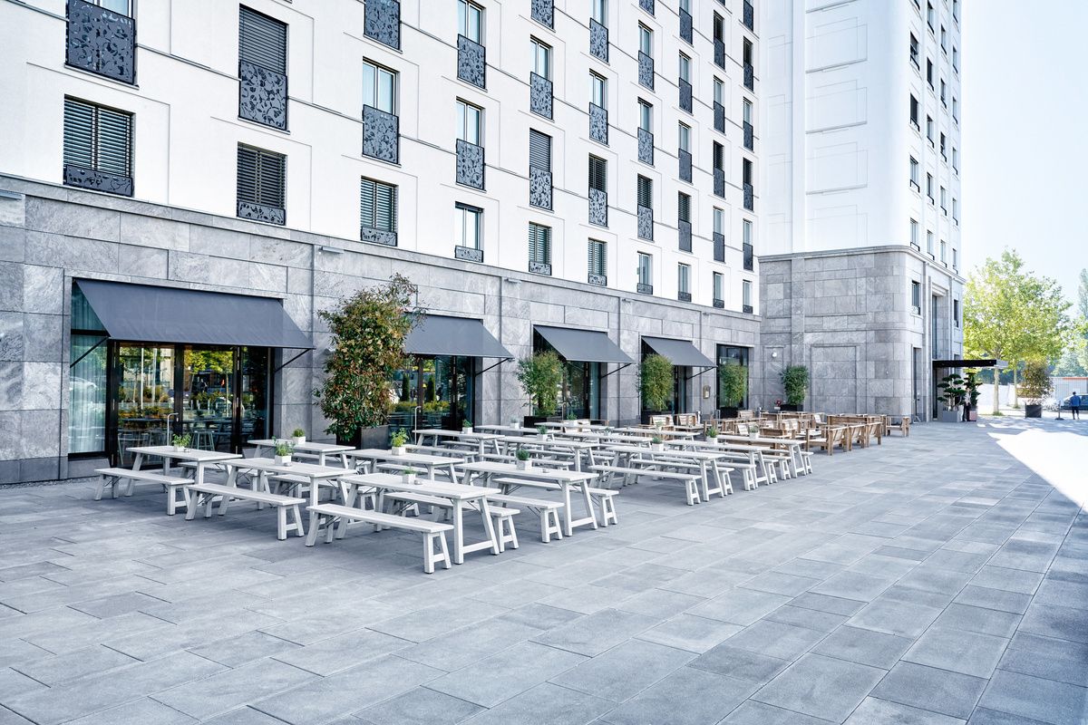 Große Außenfläche eines Hotels mit Reihen von weißen Holzbänken und Tischen, Sonnenschutz über Fenstern, ideal für entspannte Mahlzeiten oder Treffen im Freien.