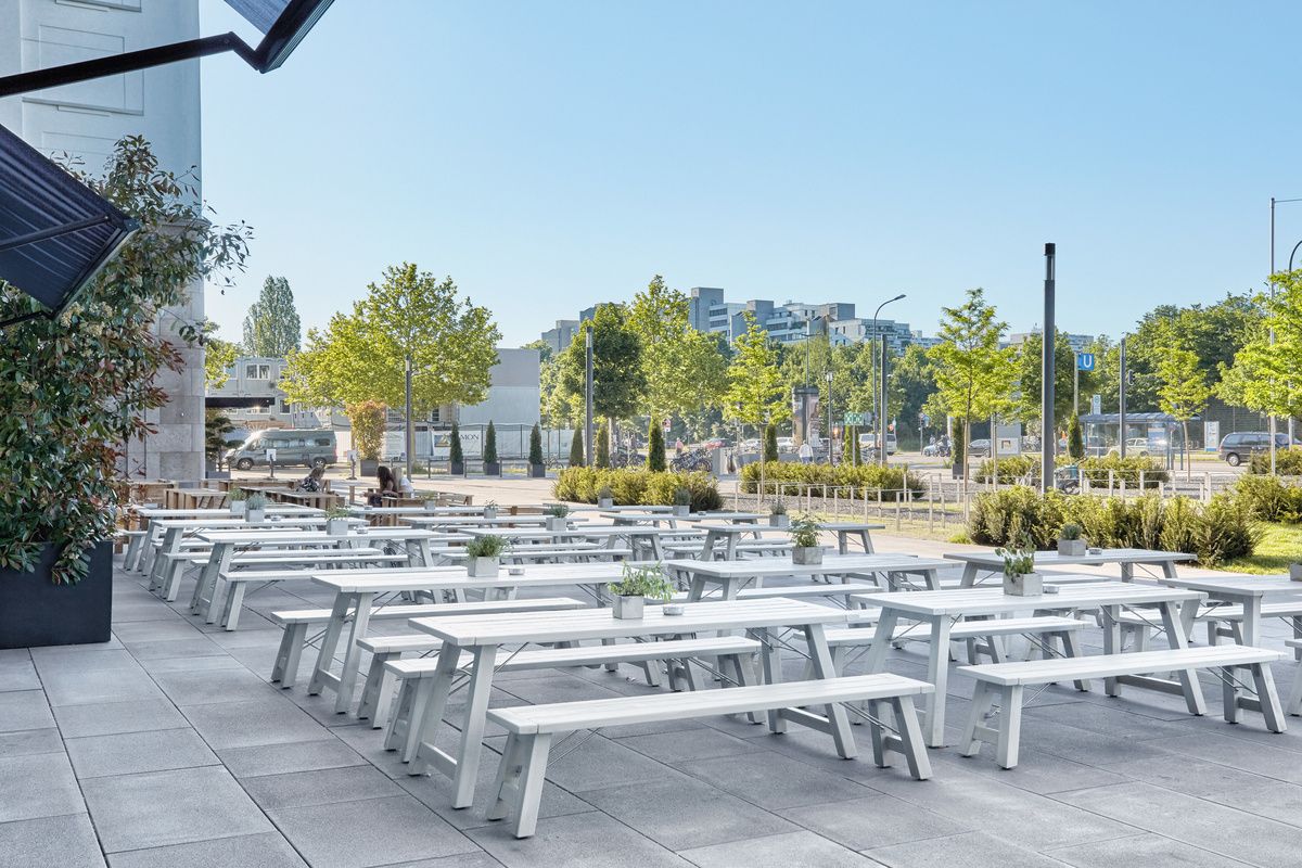 Helle, moderne Außenterrasse mit zahlreichen weißen Holzbänken und Tischen, umgeben von urbanem Grün und klarem Himmel, ideal für entspannte Pausen und geselliges Beisammensein.