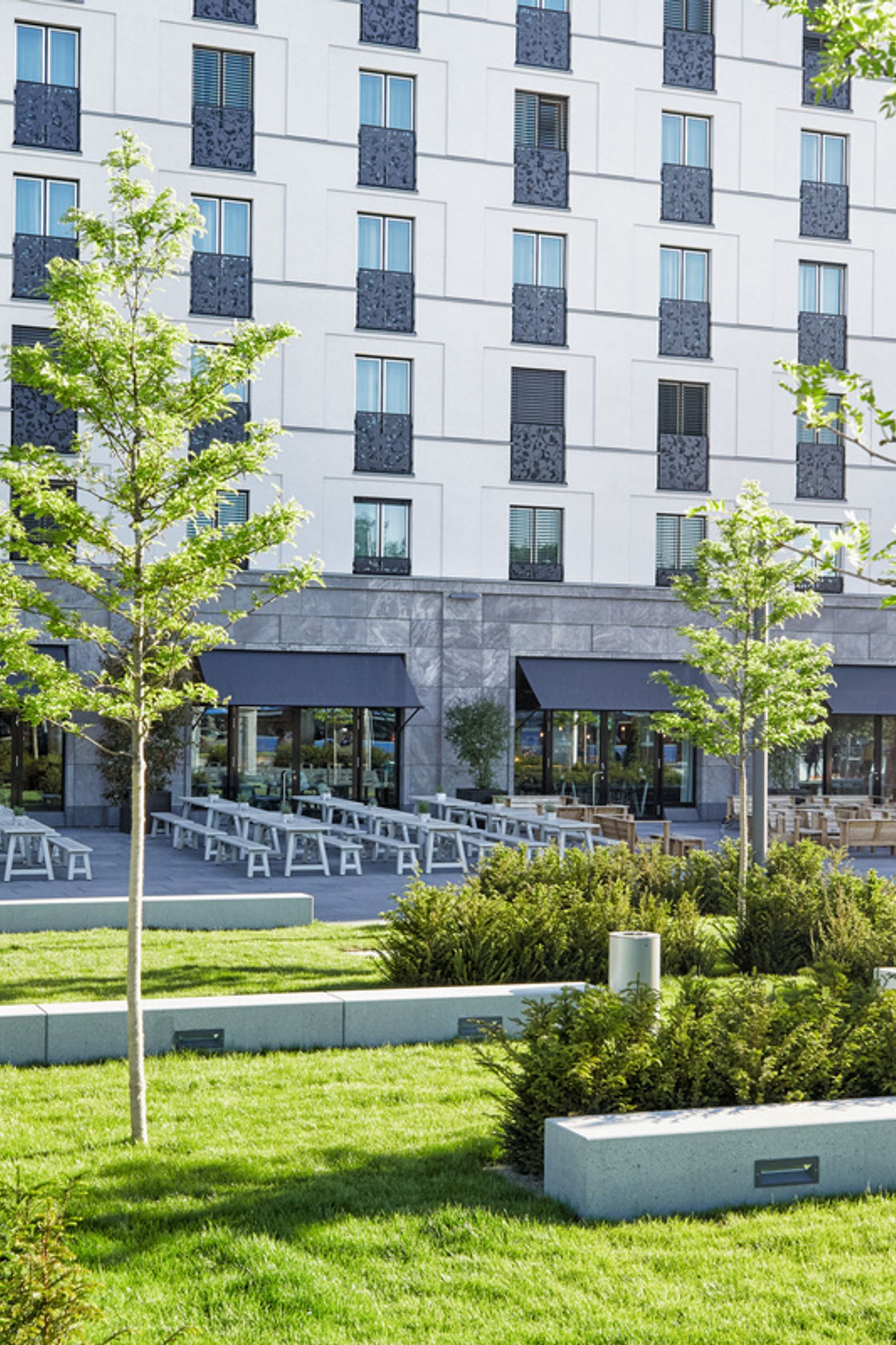 Außenansicht eines modernen Hotels mit großen Fenstern, Terrasse mit Sitzgelegenheiten und grüner, gepflegter Gartenanlage mit jungen Bäumen und Sträuchern, die zum Entspannen und Verweilen einlädt.