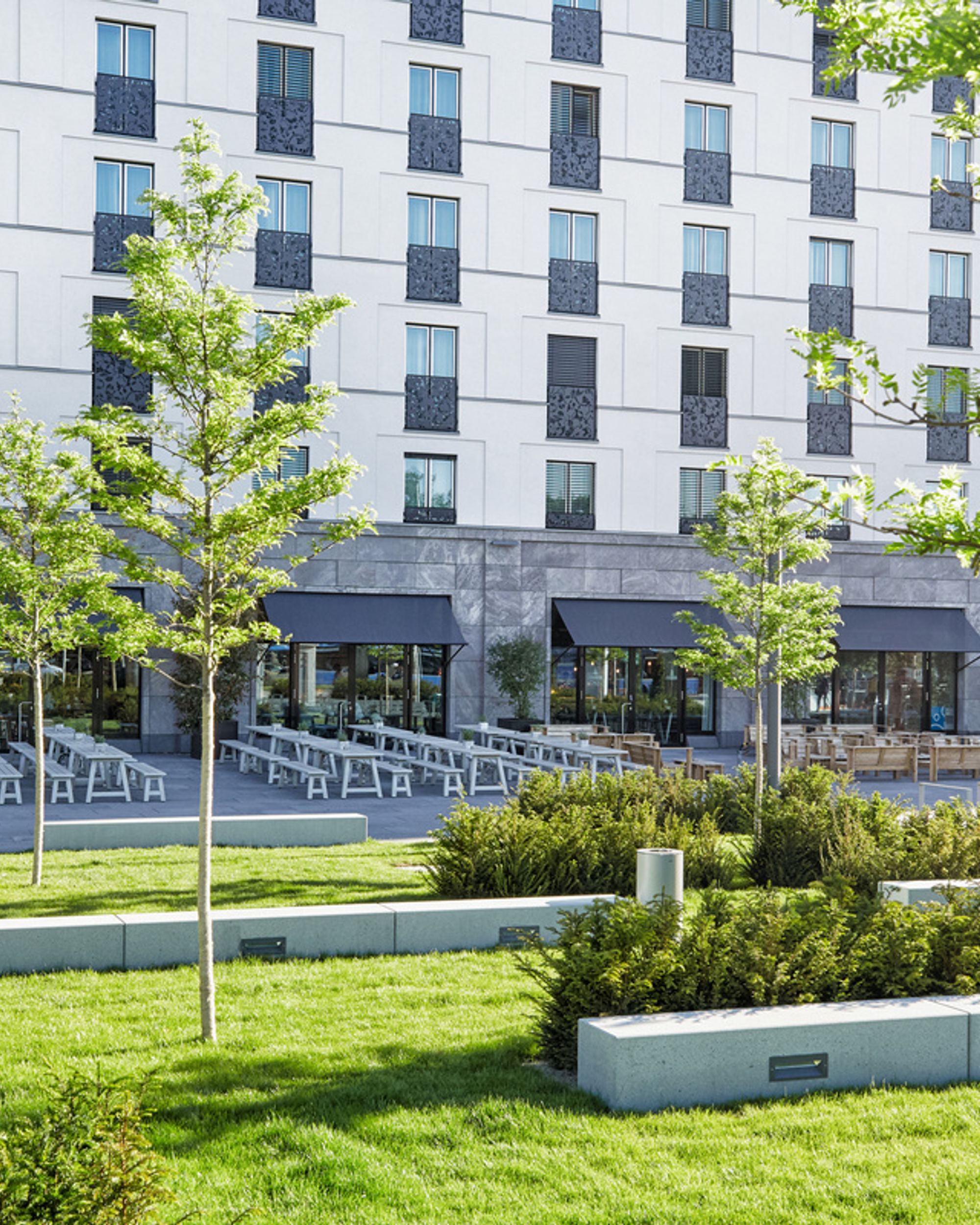 Außenansicht eines modernen Hotels mit großen Fenstern, Terrasse mit Sitzgelegenheiten und grüner, gepflegter Gartenanlage mit jungen Bäumen und Sträuchern, die zum Entspannen und Verweilen einlädt.