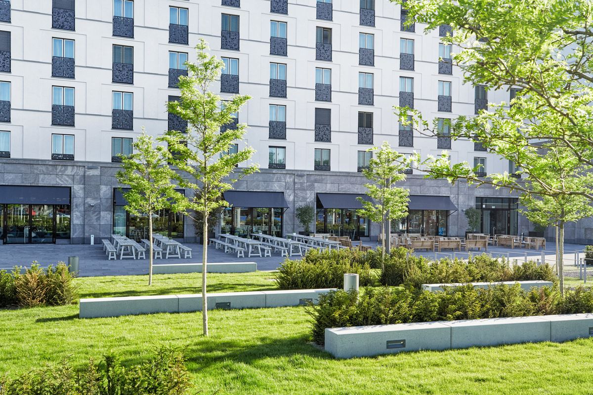 Außenansicht eines modernen Hotels mit großen Fenstern, Terrasse mit Sitzgelegenheiten und grüner, gepflegter Gartenanlage mit jungen Bäumen und Sträuchern, die zum Entspannen und Verweilen einlädt. Außenansicht eines modernen Hotels mit großen Fenstern, Terrasse mit Sitzgelegenheiten und grüner, gepflegter Gartenanlage mit jungen Bäumen und Sträuchern, die zum Entspannen und Verweilen einlädt.