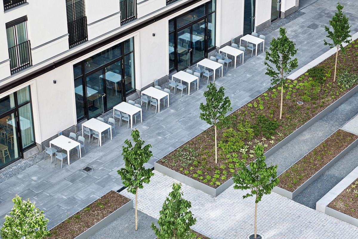 Moderne Außenterrasse mit weißen Tischen und Stühlen neben einem Hotelgebäude, umgeben von gepflegten Grünflächen mit jungen Bäumen, ideal für entspannte Mahlzeiten oder Kaffeepausen im Freien.