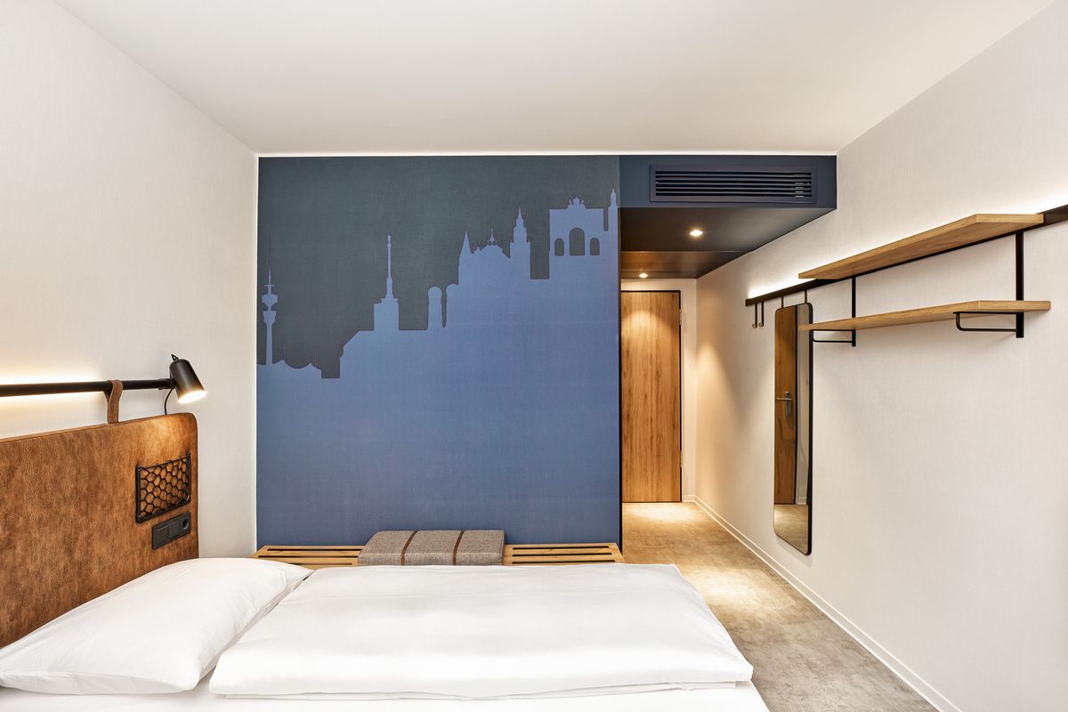 Modernes Hotelzimmer mit klarer, heller Atmosphäre, komfortablem Bett und stilvoller Wandgestaltung mit Stadtsilhouette, Holzdetails und offenem Raum für entspannte Aufenthalte.