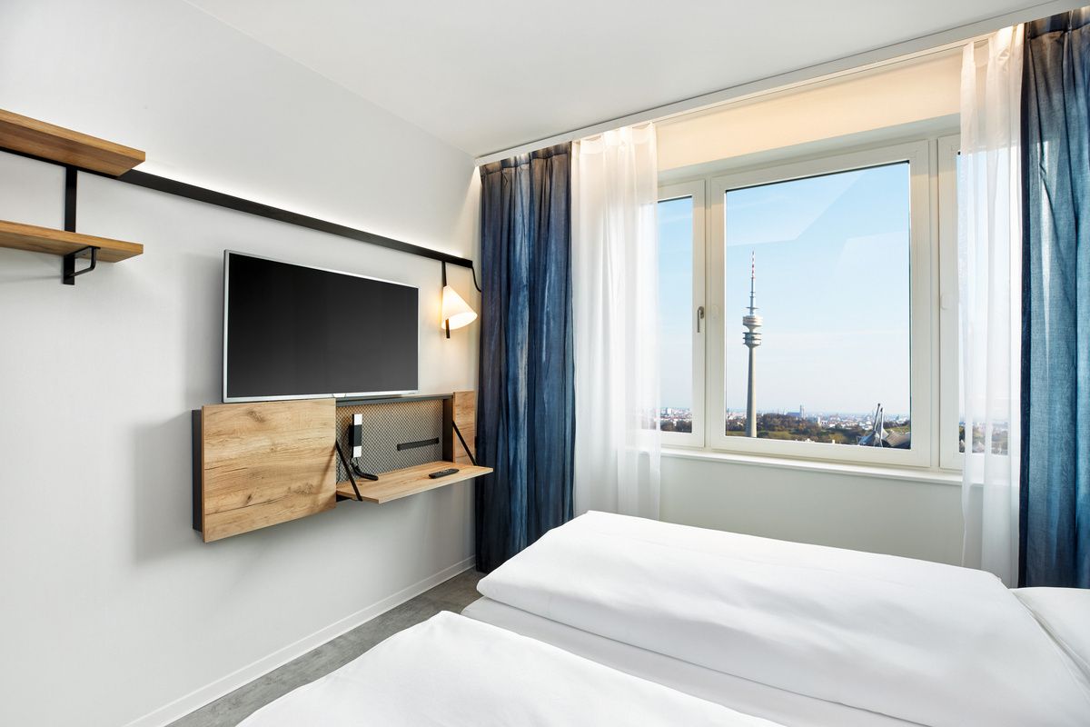 Helles, modernes Hotelzimmer mit Doppelbett, großem Fenster mit Ausblick auf Stadtturm, TV an der Wand und stilvoller Holzmöblierung, luftige blau-weiße Gardinen.
