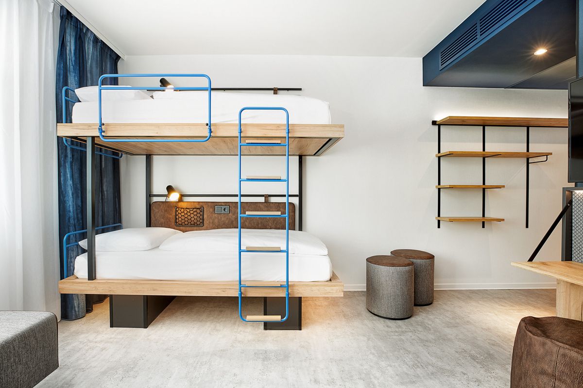 Modern eingerichtetes Hotelzimmer mit Etagenbett, minimalistischen Regalen und gemütlichen Sitzhockern, hellem Boden und neutralen Farbtönen für einen komfortablen Aufenthalt.