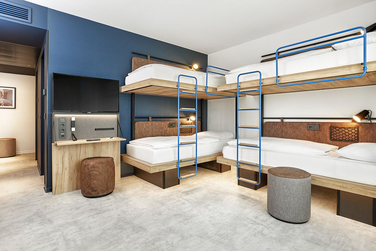 Modern eingerichtetes Mehrbettzimmer mit zwei Etagenbetten, hellem Boden, blauen Wänden und gemütlichen Sitzhockern, ideal für Gruppen oder Familienreisende, die funktionalen Komfort und eine freundliche Atmosphäre suchen.