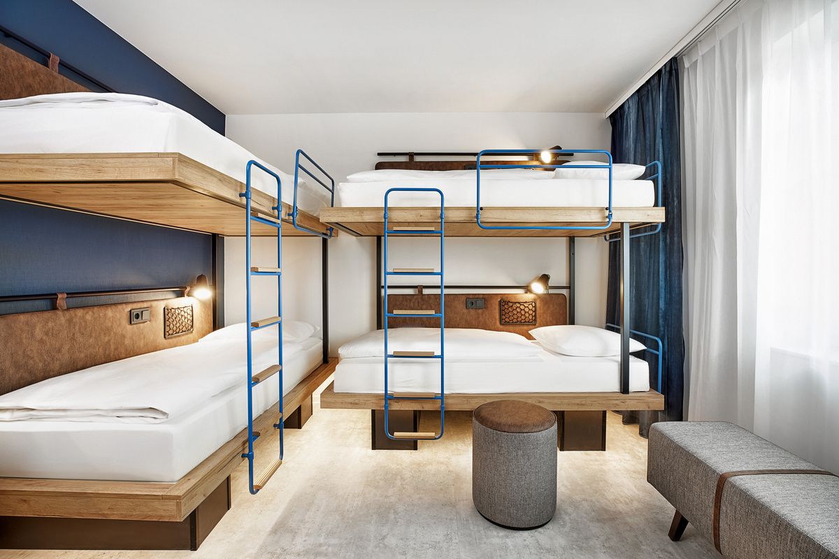 Doppelstockbetten mit modernem Design in einem hellen, freundlichen Zimmer, ausgestattet mit blauen Metallgeländern, weichen Matratzen und indirekter Beleuchtung. Vor einem großen Fenster liegen ein grauer Hocker und eine Sitzbank, ideal für entspannte Aufenthalte.