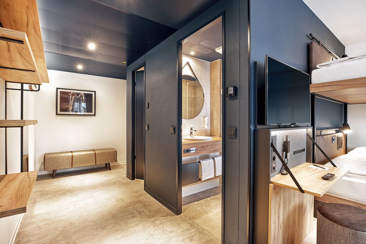 Modern eingerichtetes Hotelzimmer mit hellem Boden, geräumigem Bad, Doppelstockbett, Schreibtisch mit TV und angenehmer Beleuchtung für Komfort und Funktionalität während des Aufenthalts.
