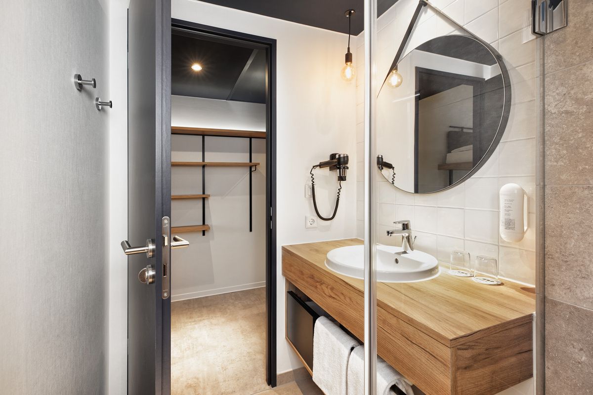 Modern eingerichtetes Badezimmer mit Holz-Waschtisch, rundem Spiegel und Haartrockner, angrenzend an begehbaren Kleiderschrank mit offenen Regalen, klare Linien und warme Beleuchtung schaffen eine einladende Atmosphäre.