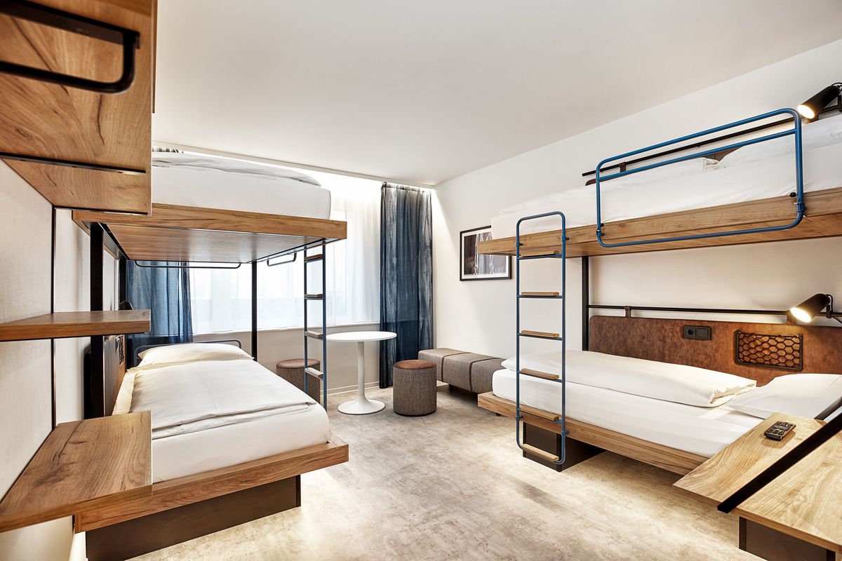 Schlicht eingerichteter Mehrbettzimmer mit zwei Etagenbetten aus Holz, hellem Boden, großem Fenster mit Vorhängen sowie Sitzgelegenheiten und kleinem Tisch, ideal für Gruppenreisende oder Familien.