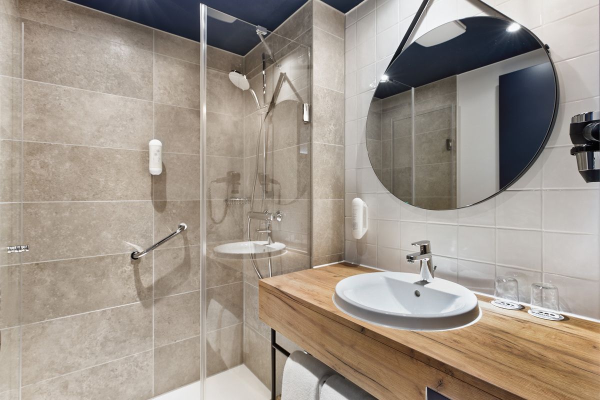 Modernes Badezimmer mit begehbarer Dusche, runder Spiegel, weißem Waschbecken auf Holzablage und hellen Fliesen. Sauber und funktional gestaltet für komfortablen Aufenthalt.