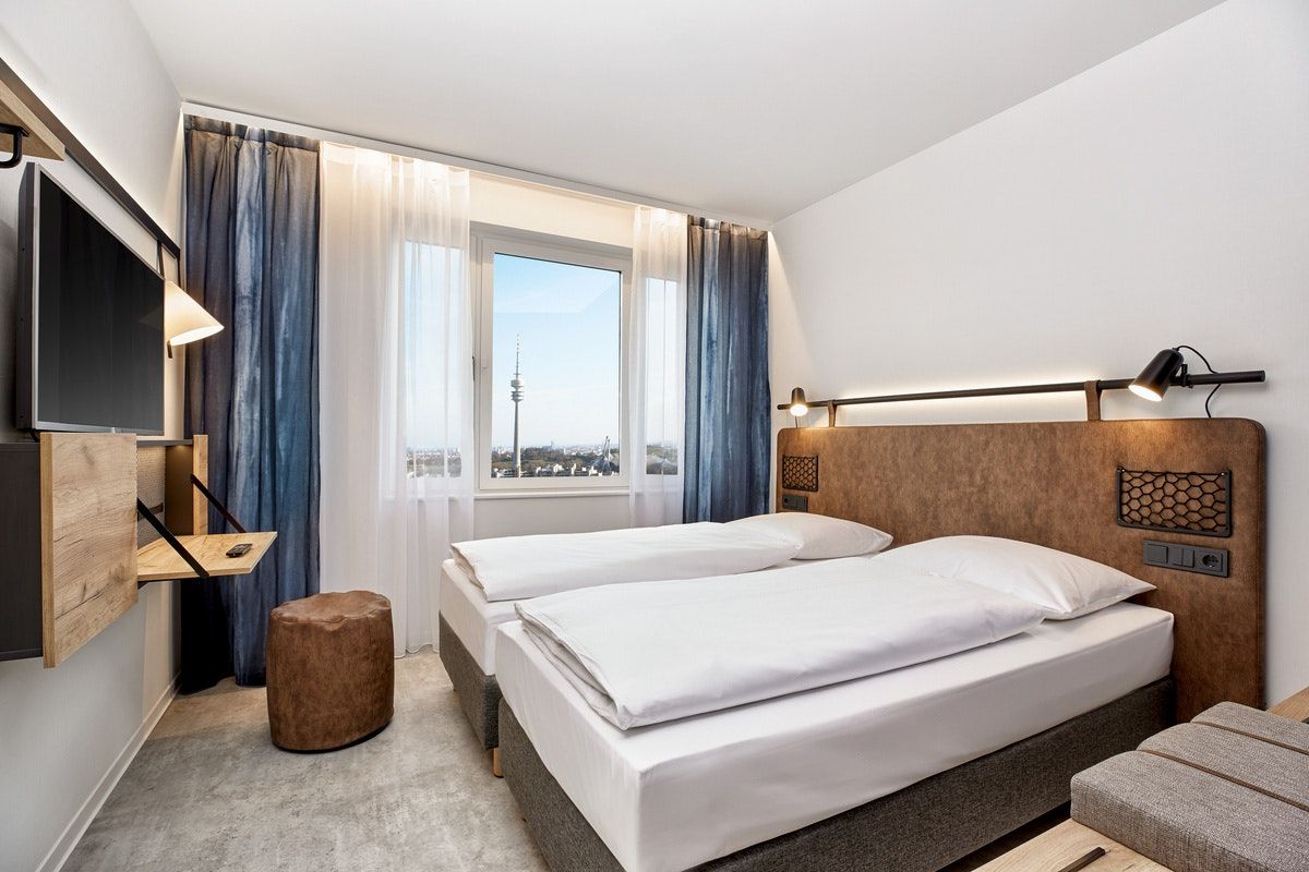 Modernes, helles Hotelzimmer mit zwei Einzelbetten, großem Fenster mit Blick auf eine Stadt und Fernseher an der Wand. Das Zimmer wirkt ruhig und einladend, ideal für Geschäftsreisende oder Urlaubsgäste.