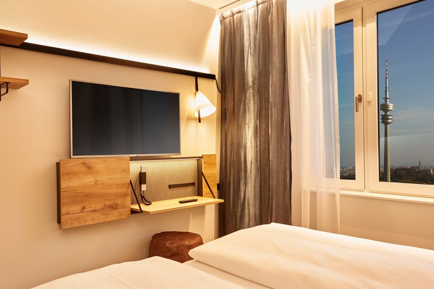 Helles Hotelzimmer mit zwei Betten, großem Fenster und Blick auf Fernsehturm. Moderne Einrichtung mit Wand-TV, Schreibtisch und gemütlicher Atmosphäre.
