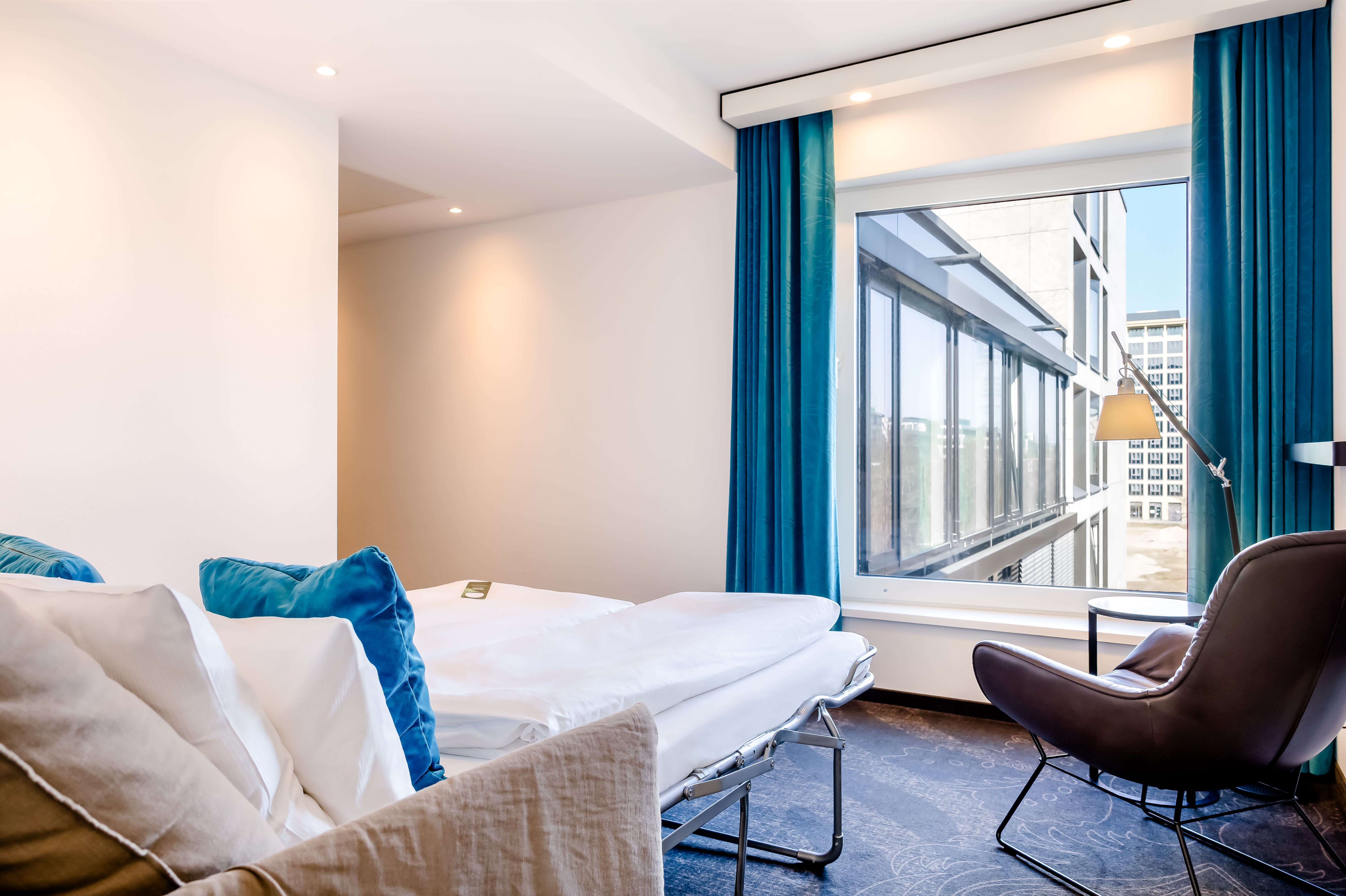Hell eingerichtetes Hotelzimmer mit gemütlichem Bett, blauen Vorhängen und einem stilvollen Sessel am Fenster mit Stadtblick. Ruhige, moderne Atmosphäre ideal zum Entspannen oder Arbeiten.